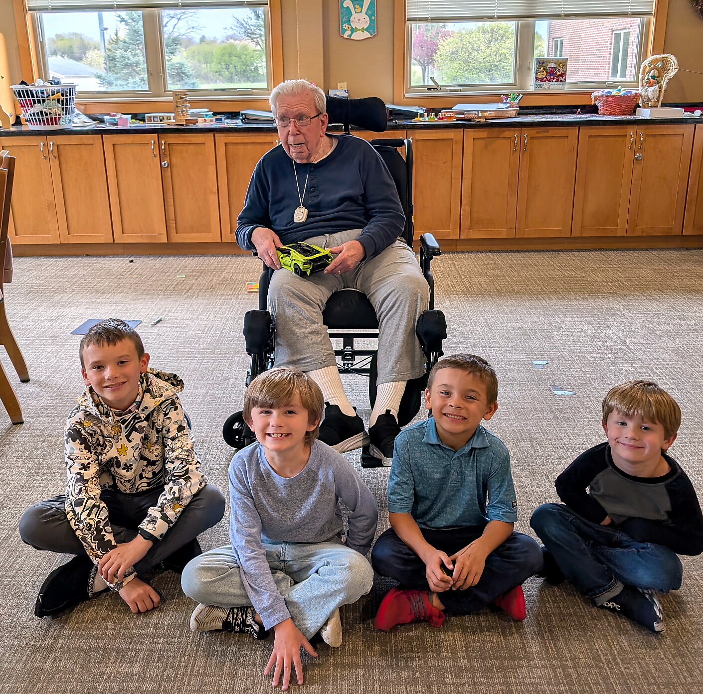 An elderly man in a wheelchair sits in a room with five smiling children on the floor in front of him, holding a bright green toy vehicle.