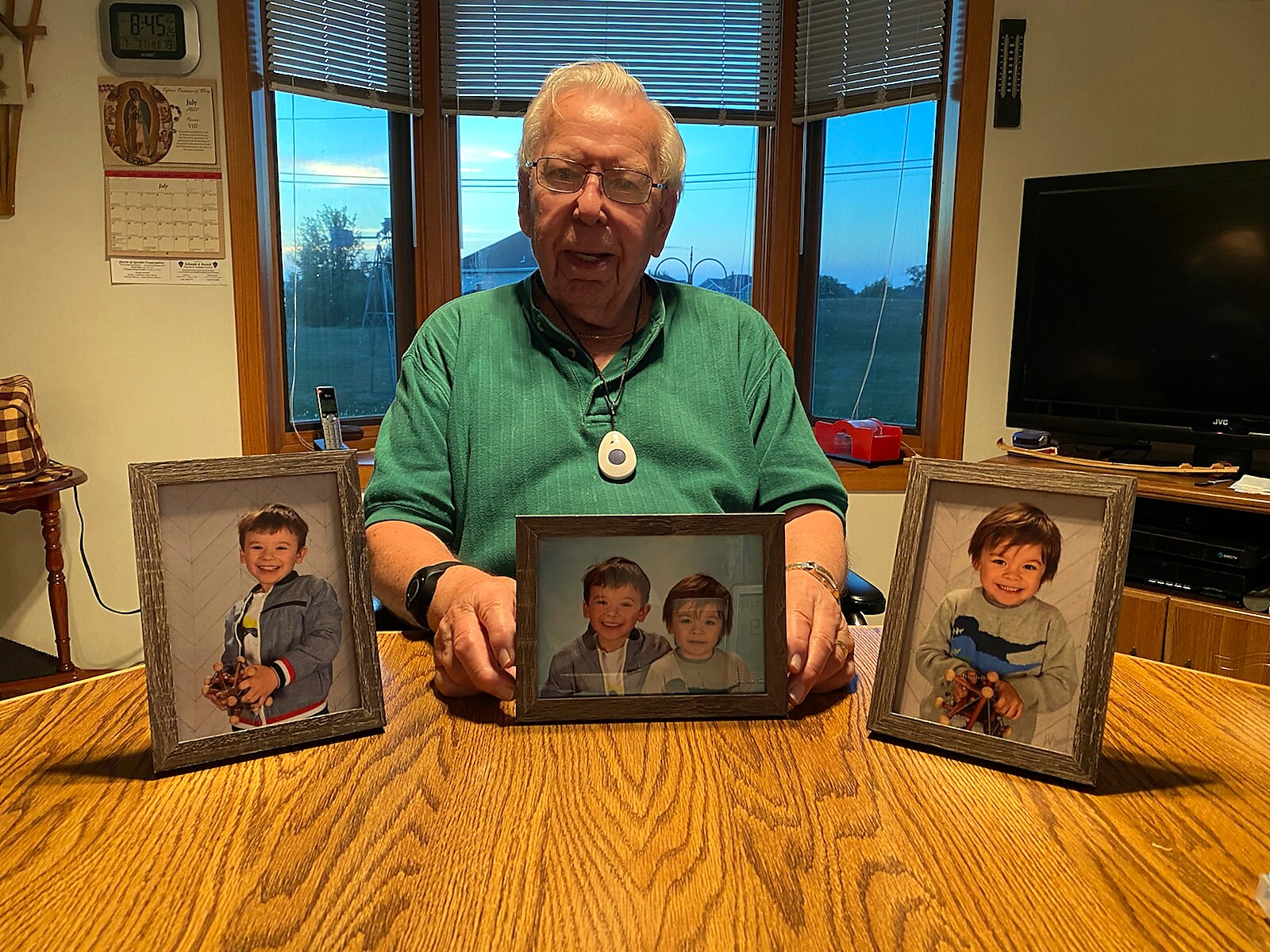 Elderly man in a green shirt sits at a wooden table with three framed photos of smiling children in front of him.