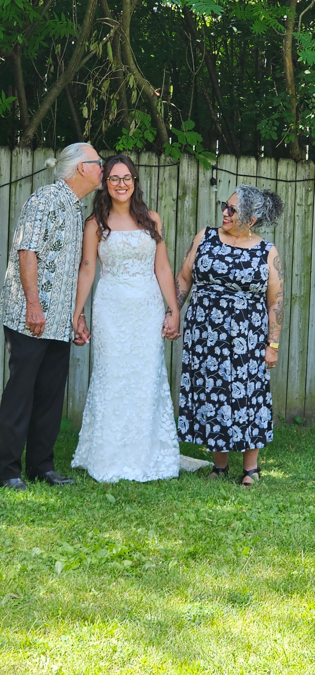 Three generations posing together outdoors: a man in a patterned shirt kissing a smiling bride while holding hands with an older woman in a black floral dress by a wooden fence.