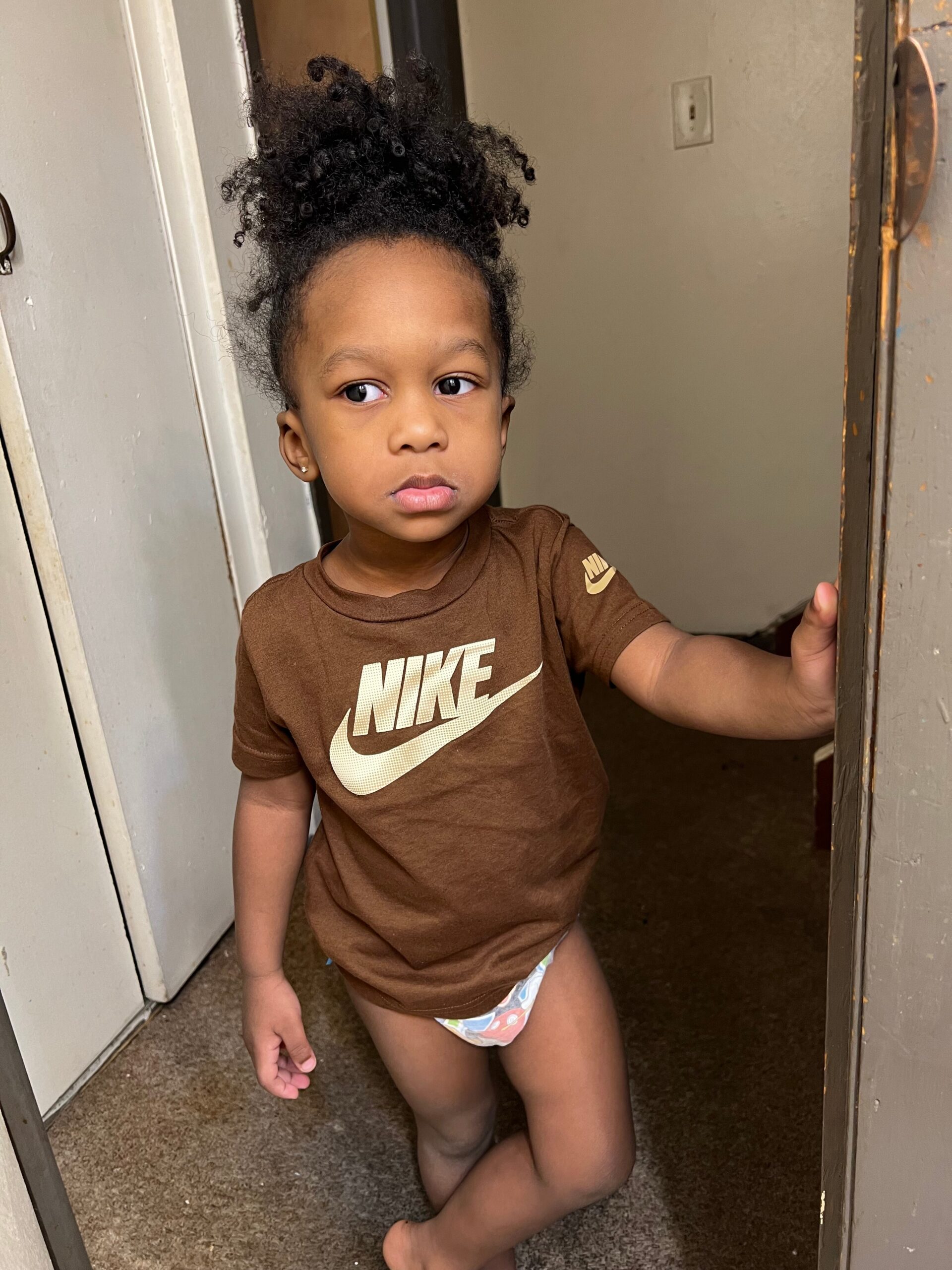 Young child standing in a doorway, wearing a brown Nike shirt and diaper, looking to the side with hair in a high puff.
