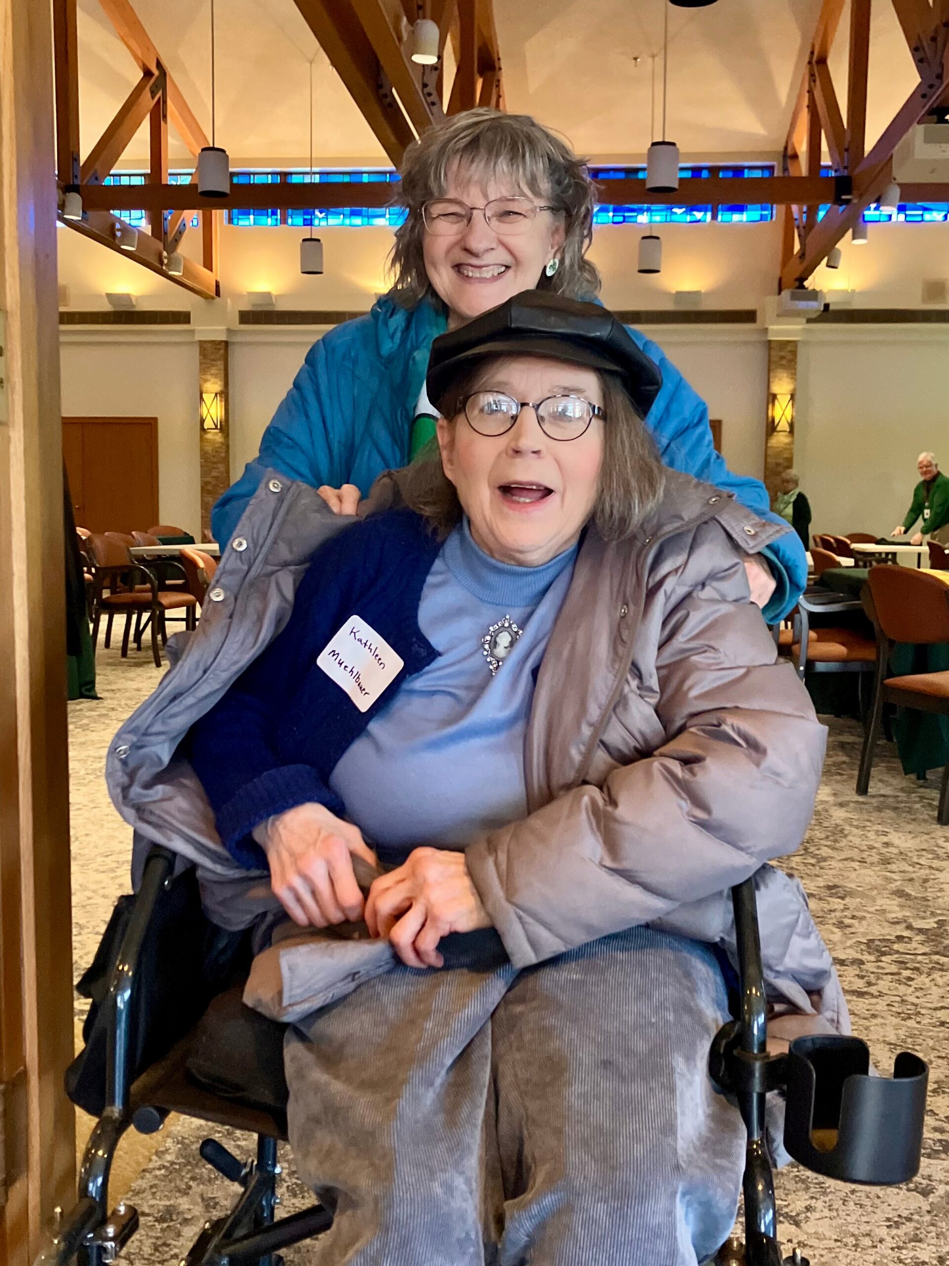 Smiling woman in a wheelchair wearing glasses and a blue top, with another woman standing behind her in a blue jacket in a sunny indoor hall.