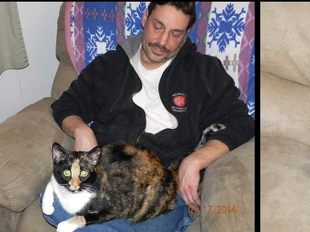 Man with mustache sits on a beige couch, wearing a white shirt and black jacket, holding a calico cat on his lap; date stamp 01/17/2014.