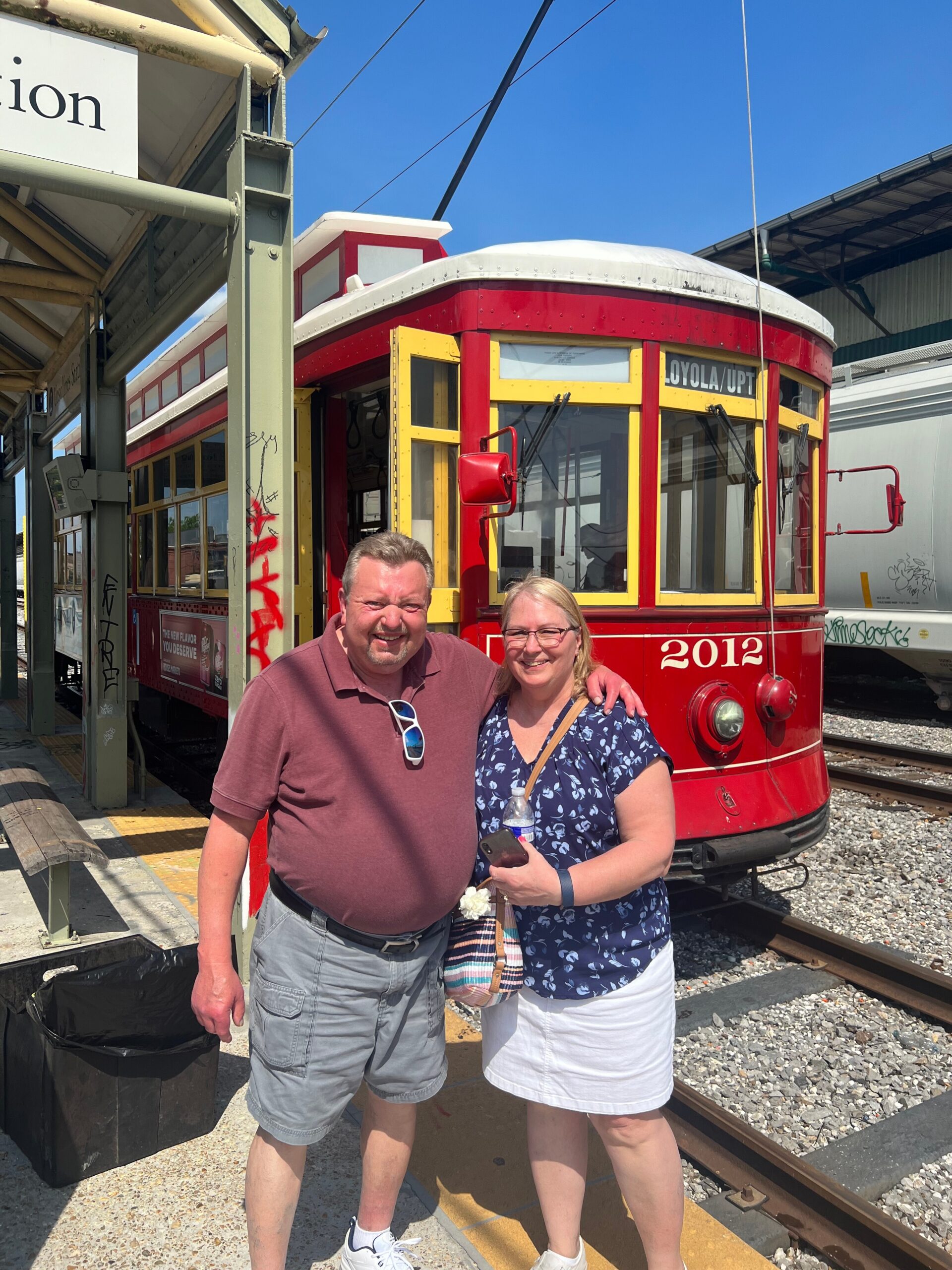 Smiling couple posing in front of a red and yellow trolley car at a station, with tracks and a clear blue sky in the background.