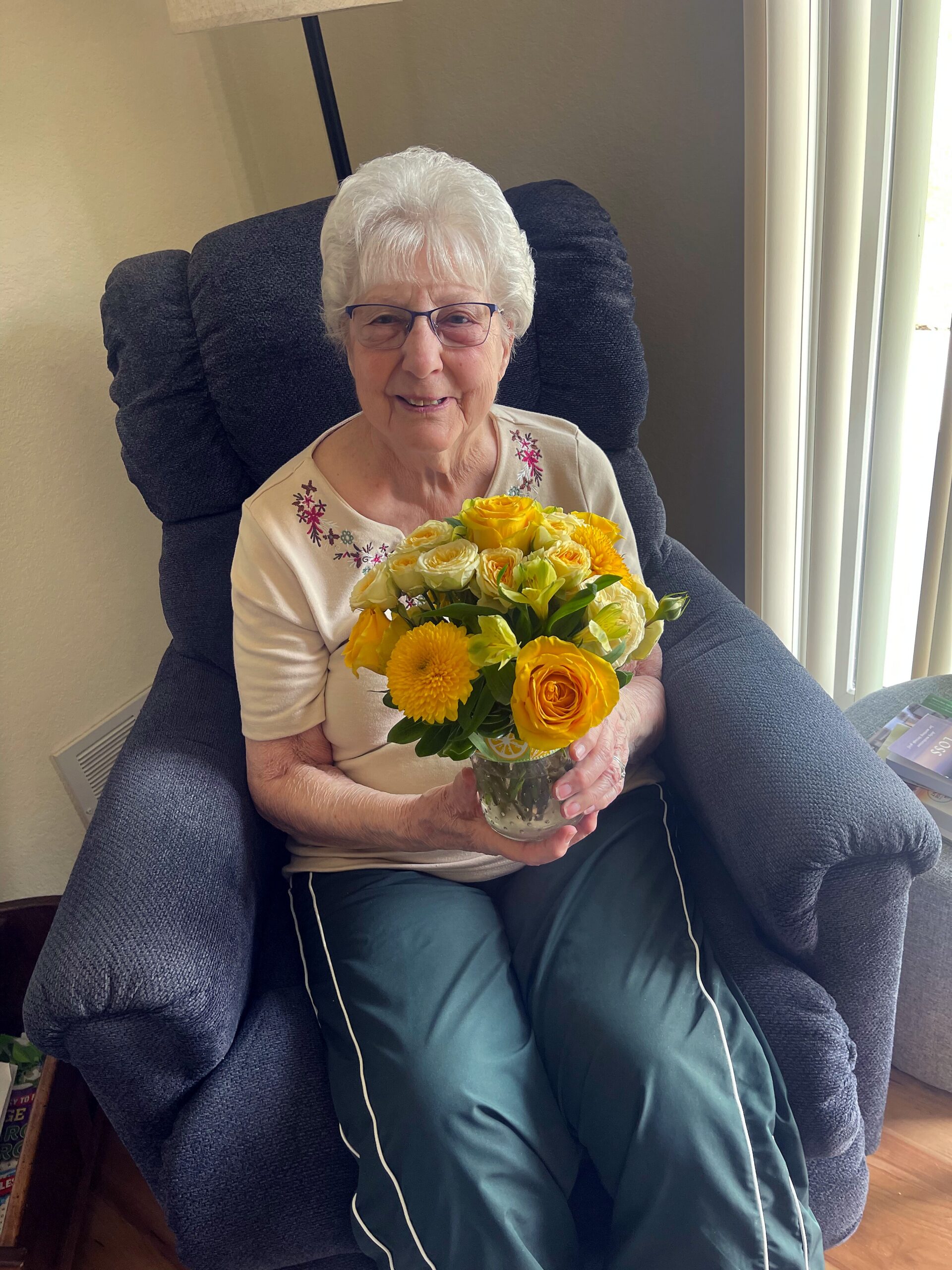 Older woman with white hair and glasses sits in a blue armchair, holding a yellow bouquet near her chest—sunlit window beside.