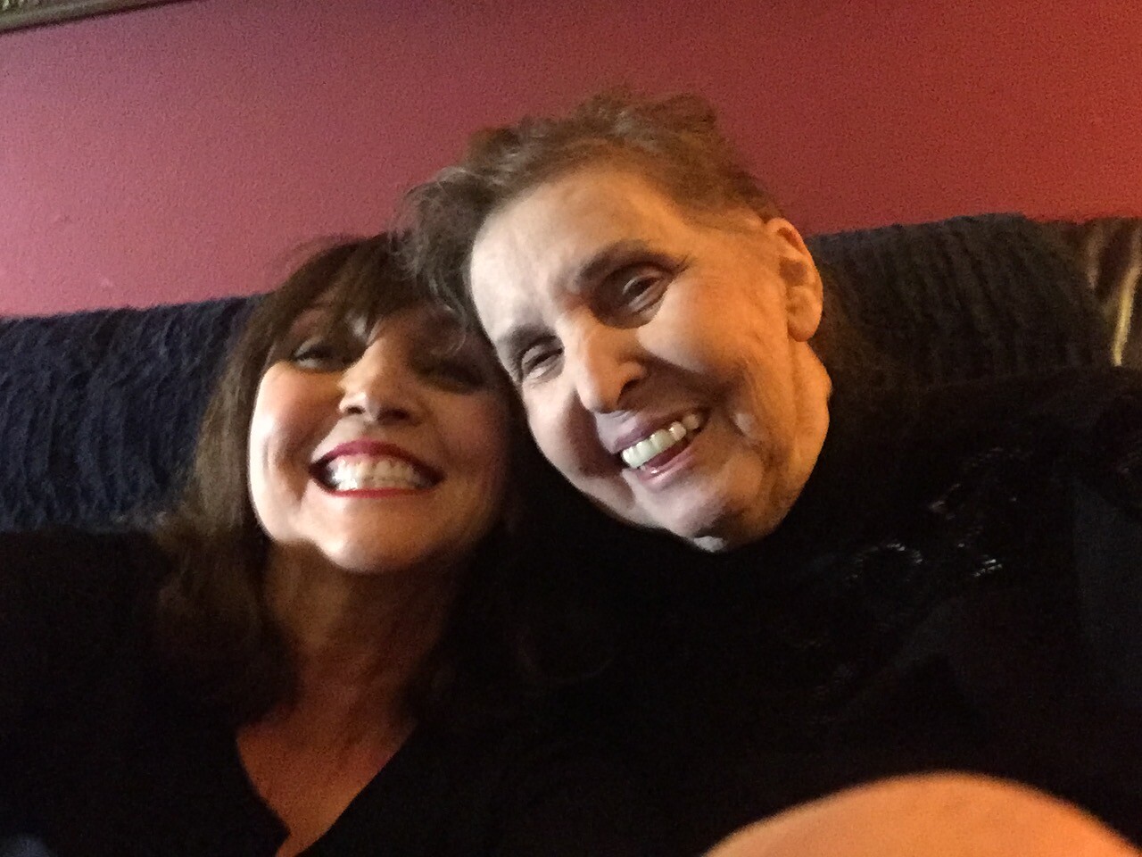 Two people smiling for a close-up selfie, woman on the left and an older man on the right, sitting together on a dark sofa with a pink wall in the background.