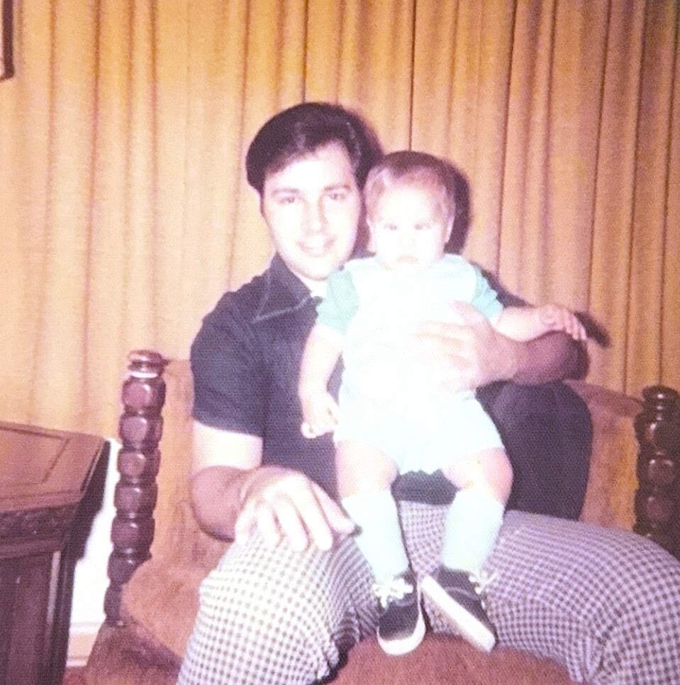 Man sitting on a couch holding a toddler on his lap, with golden curtains in the background.