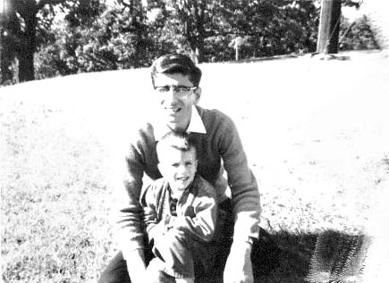 Black-and-white photo of a man kneeling with a young child on a grassy outdoor field, smiling at the camera.