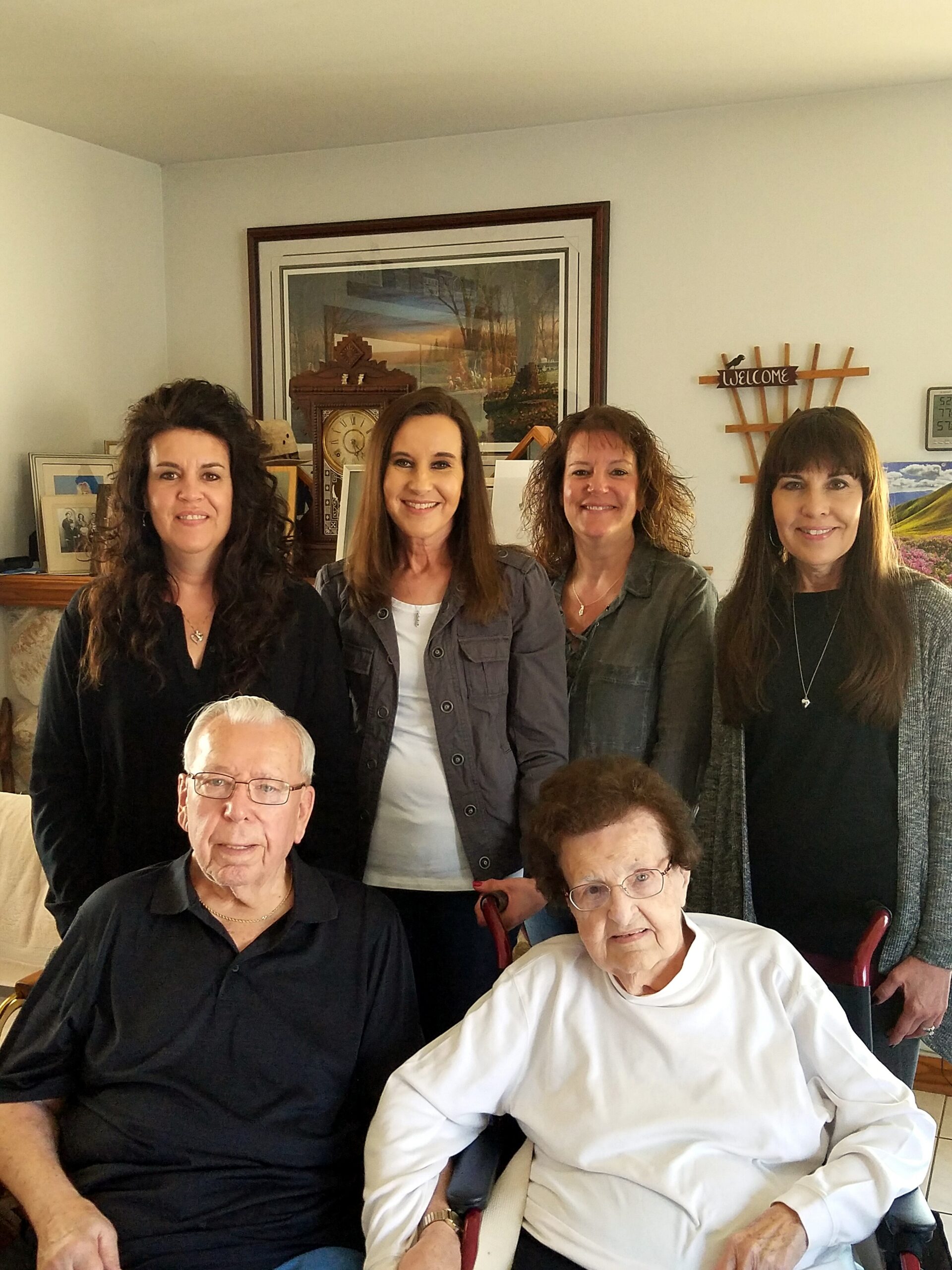 Four women stand behind two seated seniors in a living room, smiling for the photo.