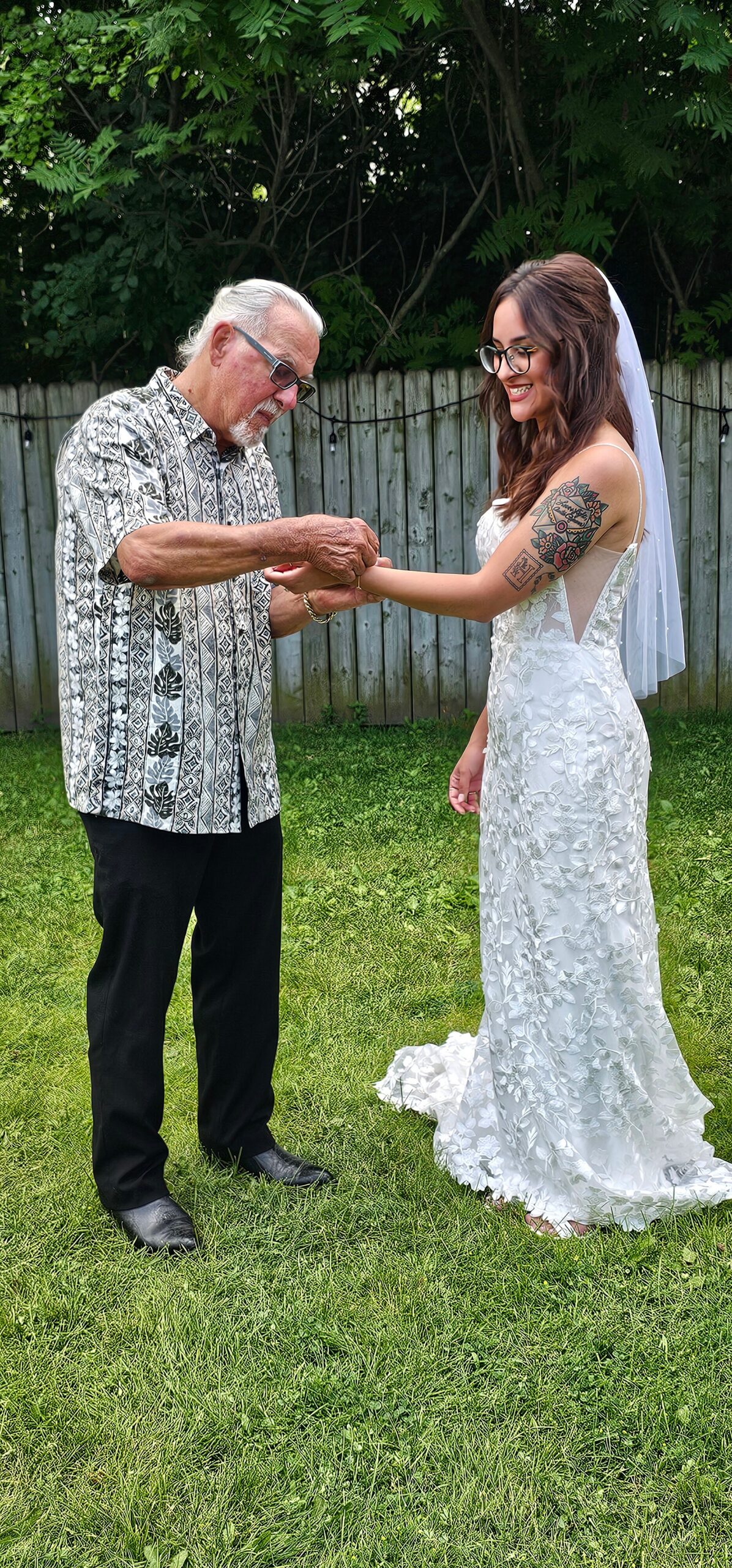 Older man in a patterned shirt helps the bride with a bracelet outdoors, a backyard setting behind them.