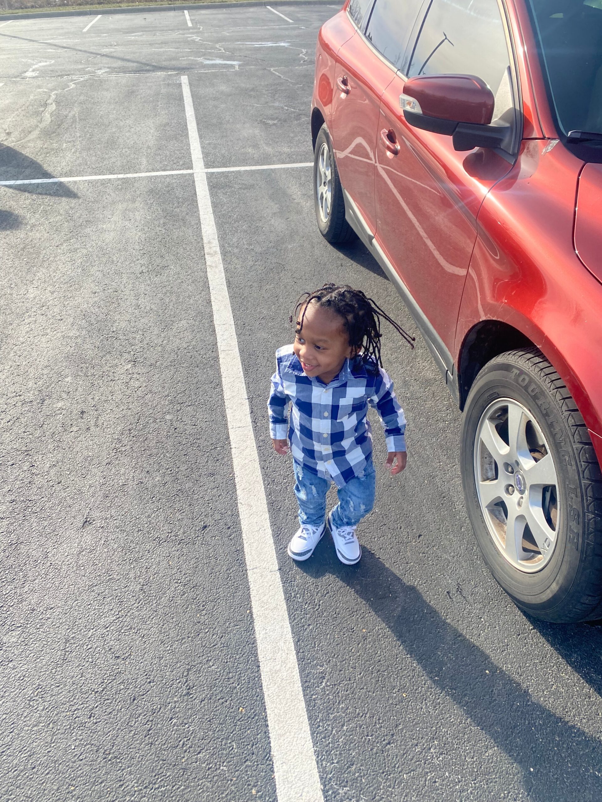 Young child in a blue and white plaid shirt standing in a parking lot beside a red SUV photographed from above.