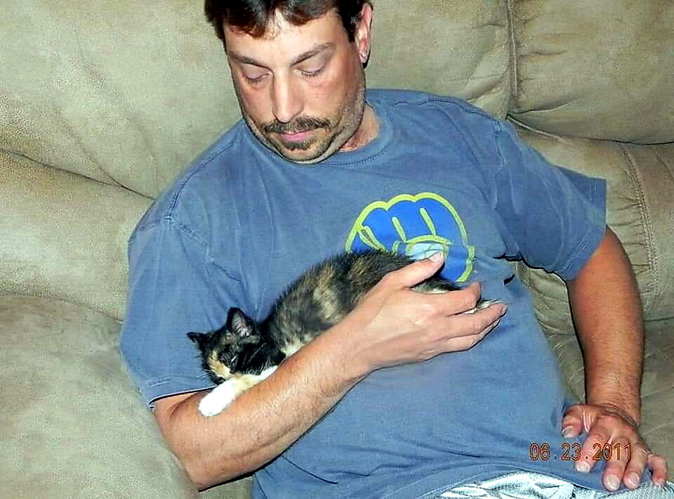 Man wearing a blue T-shirt sits on a couch, holding a small calico kitten in his arms.
