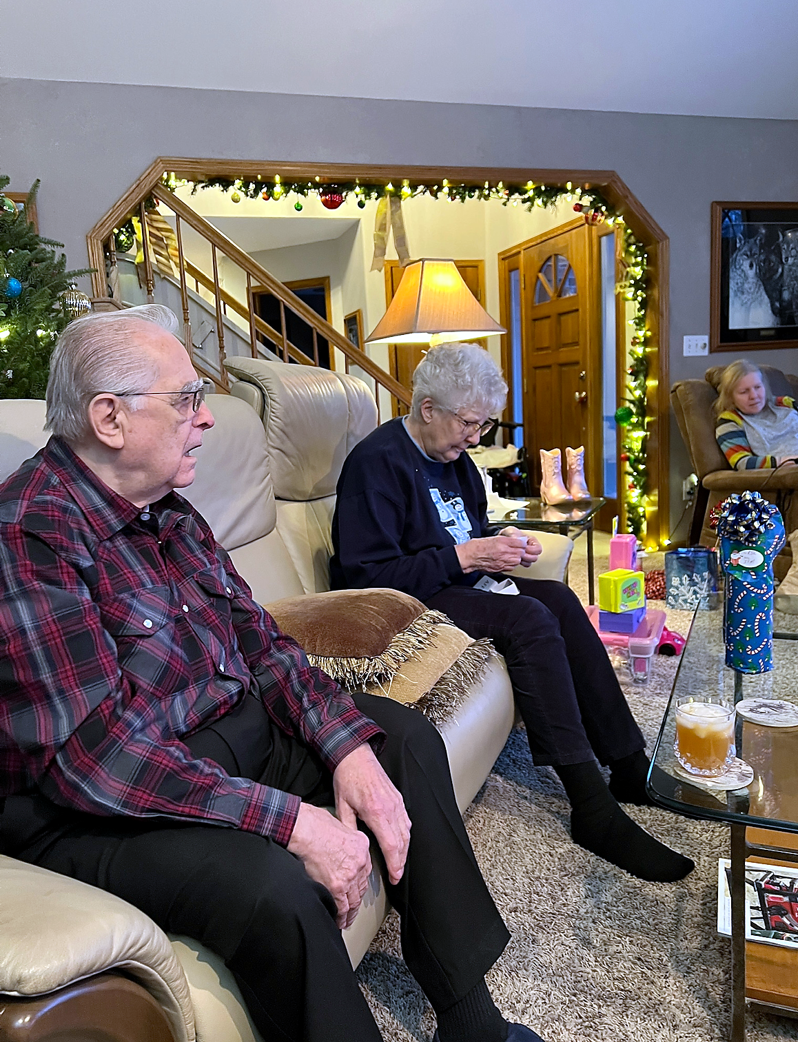 Three elderly people sit in a festively decorated living room with a Christmas tree and garlands.