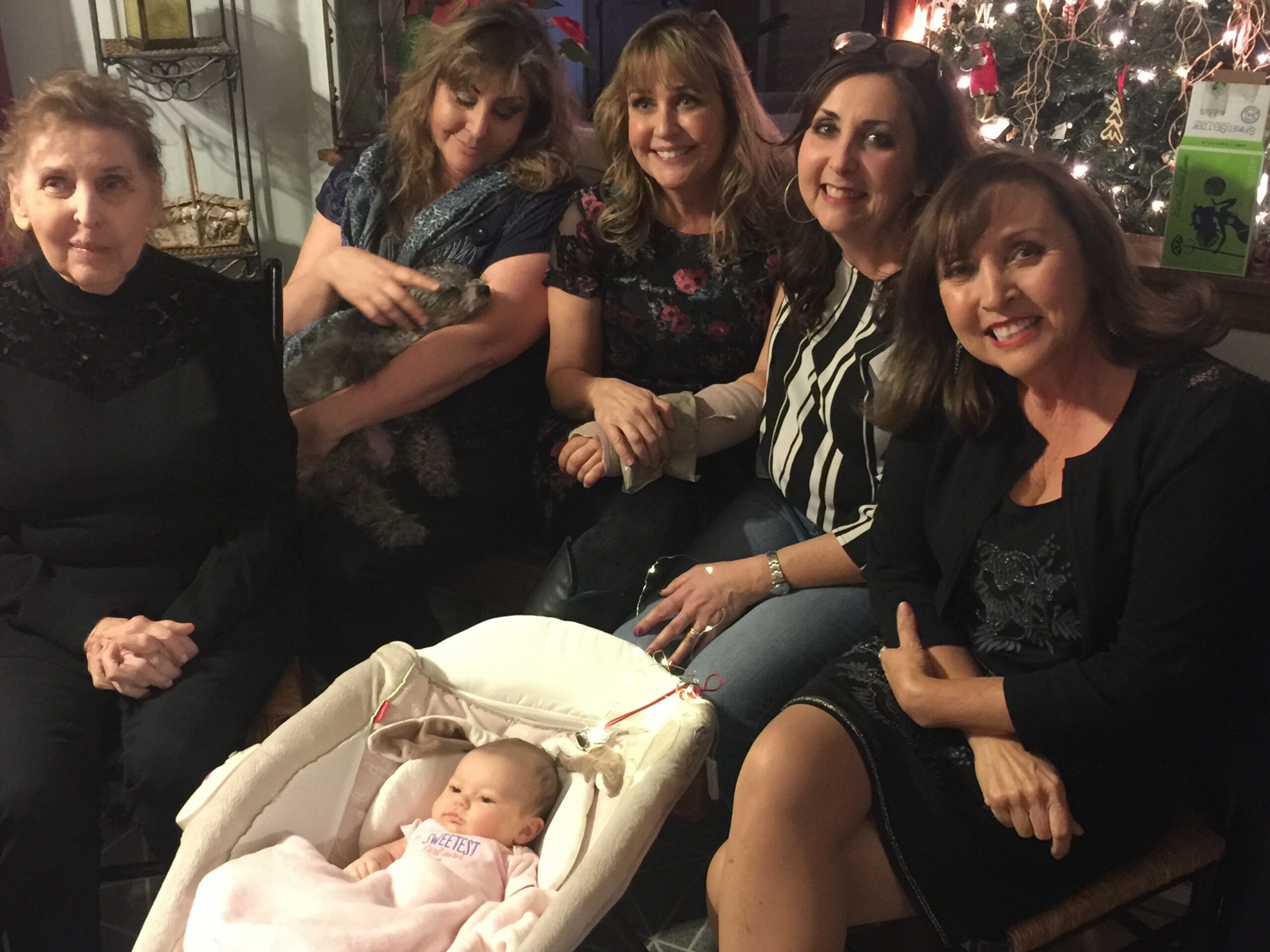 Five women sit closely together indoors, smiling, with a newborn in a bassinet in front and a Christmas tree in the background.
