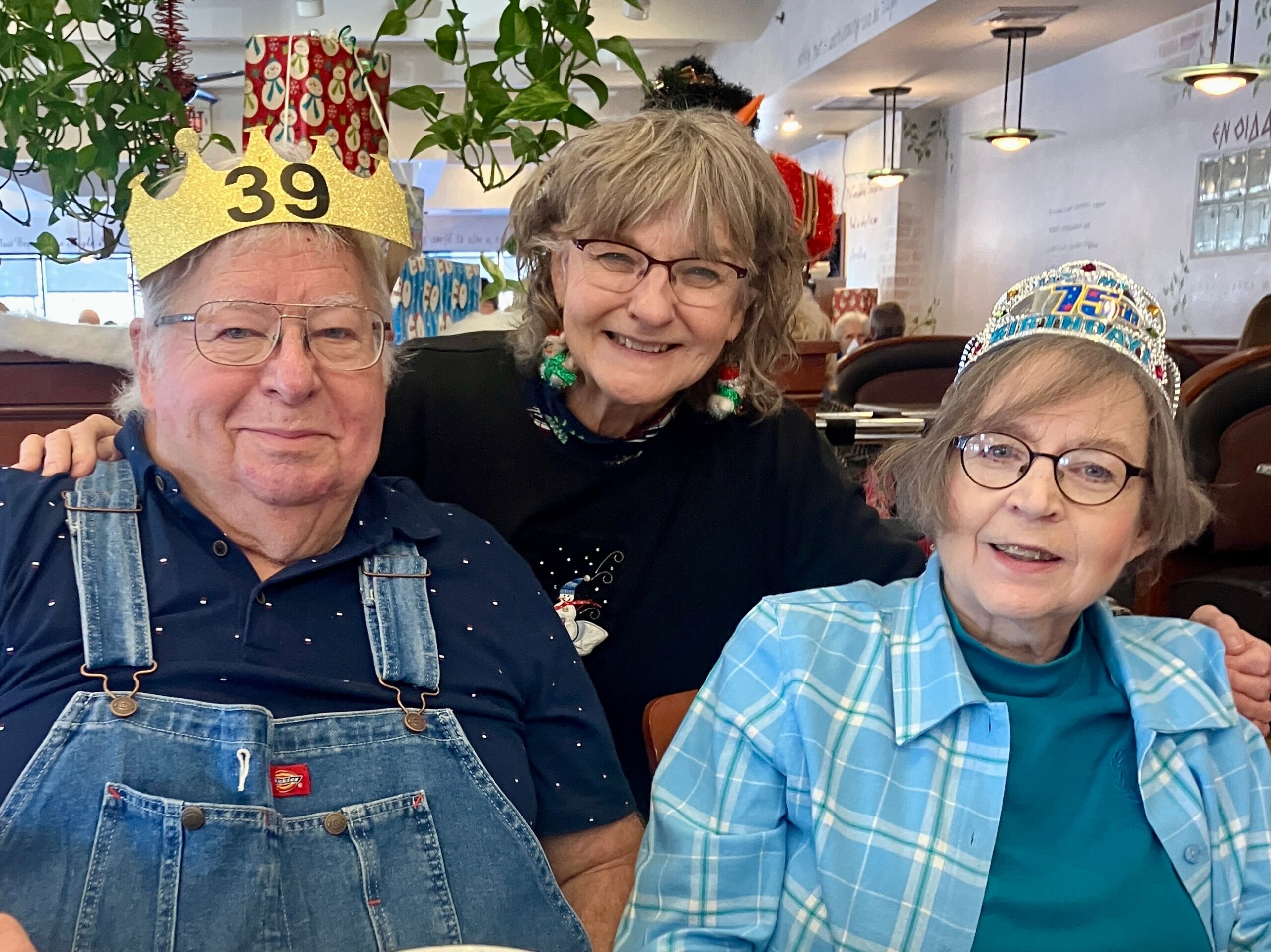 Three seniors pose for a photo at a celebration, the man on the left wearing a gold crown reading '39'. The woman in the middle smiles, wearing festive accessories; the person on the right wears a jeweled crown with '75'.