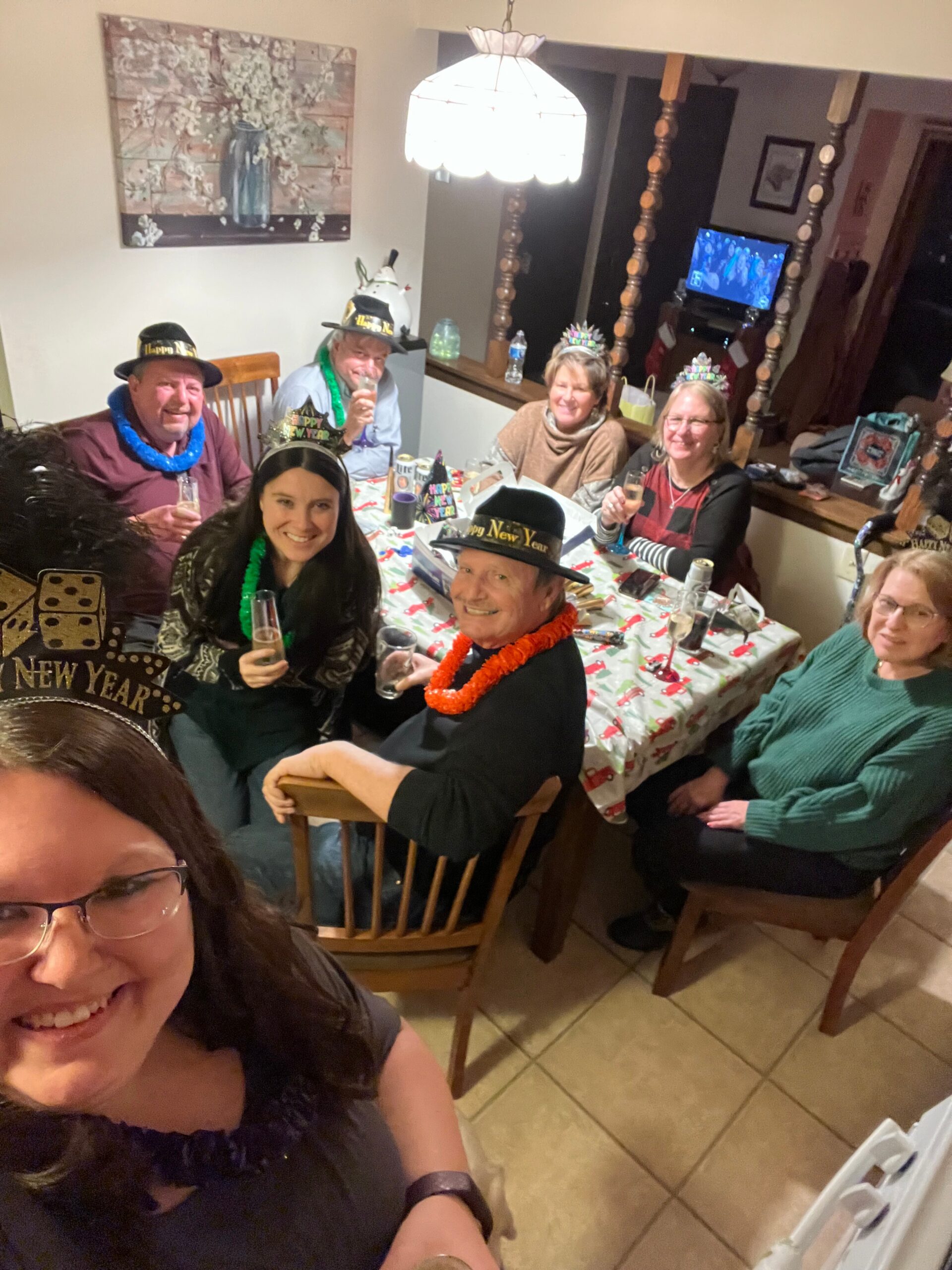 Group of adults around a dining table celebrating New Year, wearing party hats and leis, toasting with drinks.