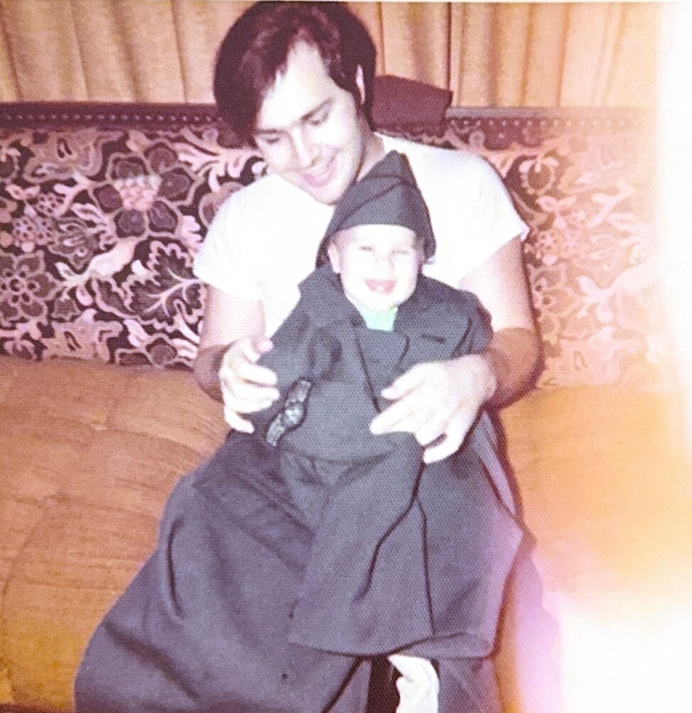 Child dressed in a black ninja costume sits on a man's lap as they smile together on a patterned couch background.