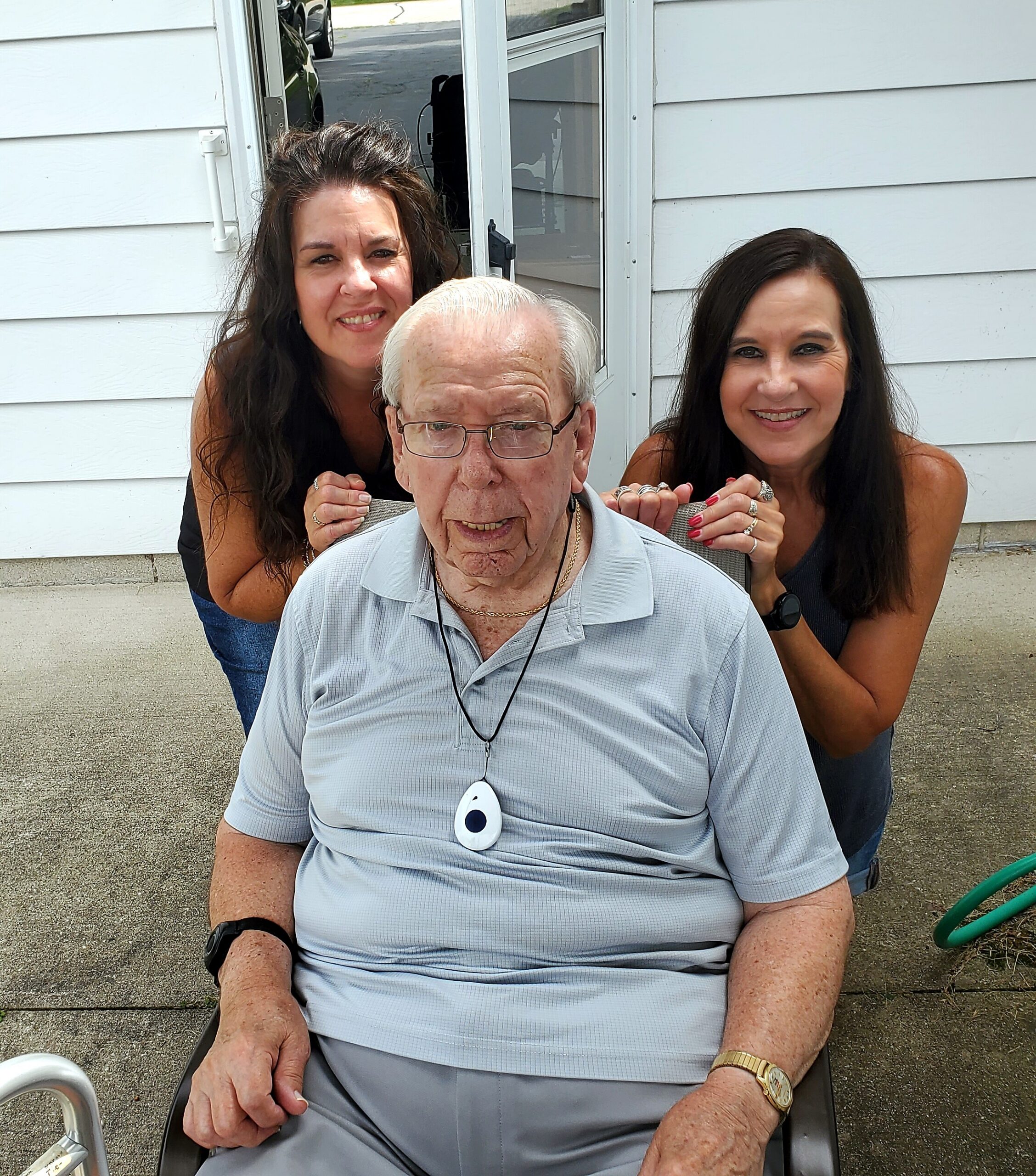 Smiling elderly man in a light blue polo sits in a wheelchair, with two women behind him outdoors near a light-gray house.