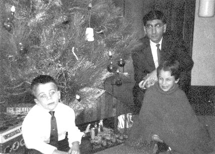 Man in a suit stands behind two children seated on the floor by a decorated Christmas tree with gifts nearby.