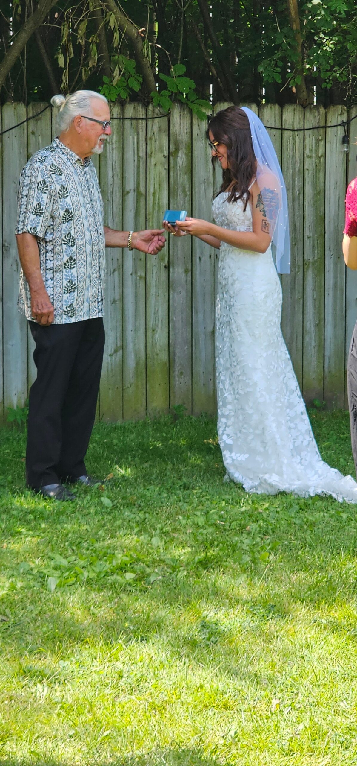 Bride in a lace wedding gown with a veil hands a small blue ring box to an older man in a patterned shirt in a backyard.