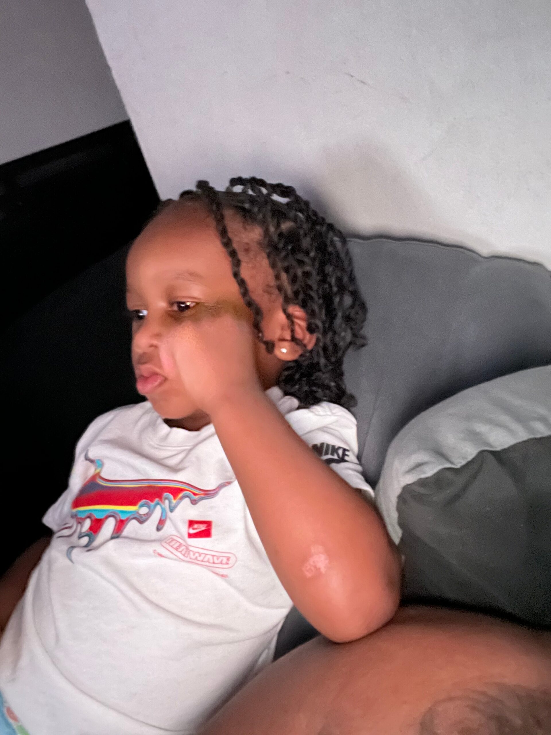 Child with braided hair lying on a bed, wearing a white graphic tee, face turned to the side on a gray pillow/blanket backdrop.