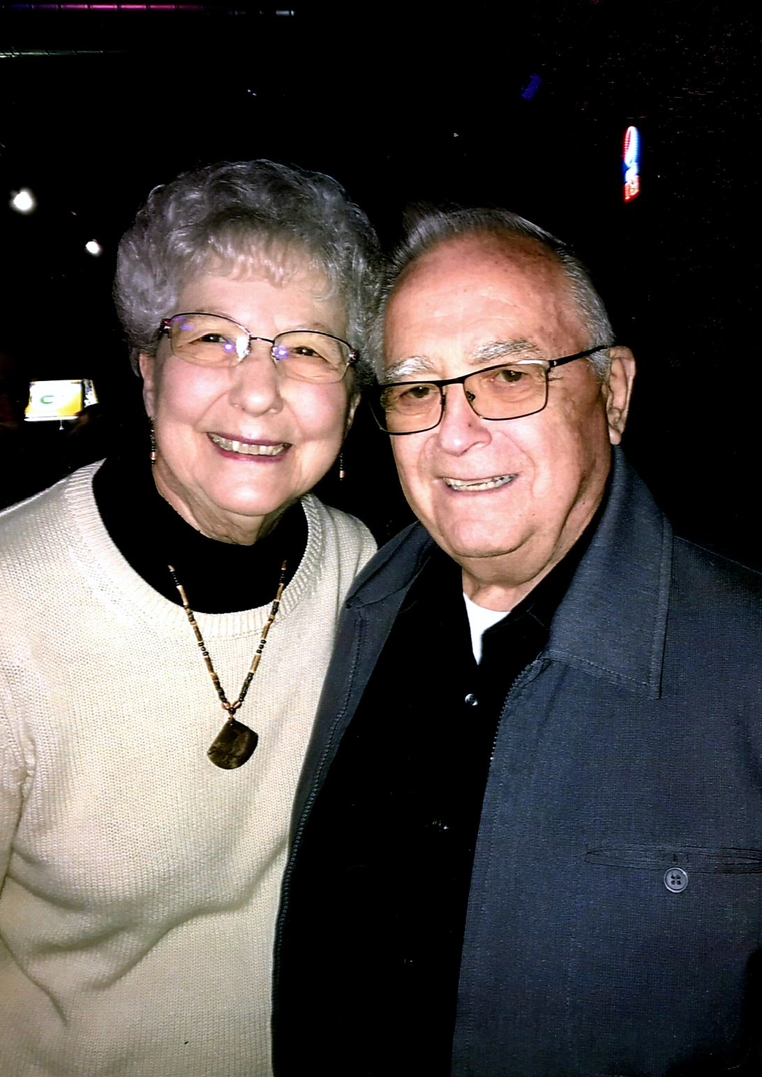 Smiling elderly woman and man pose closely for a photo at night, both wearing glasses and dark clothing with light sweaters.