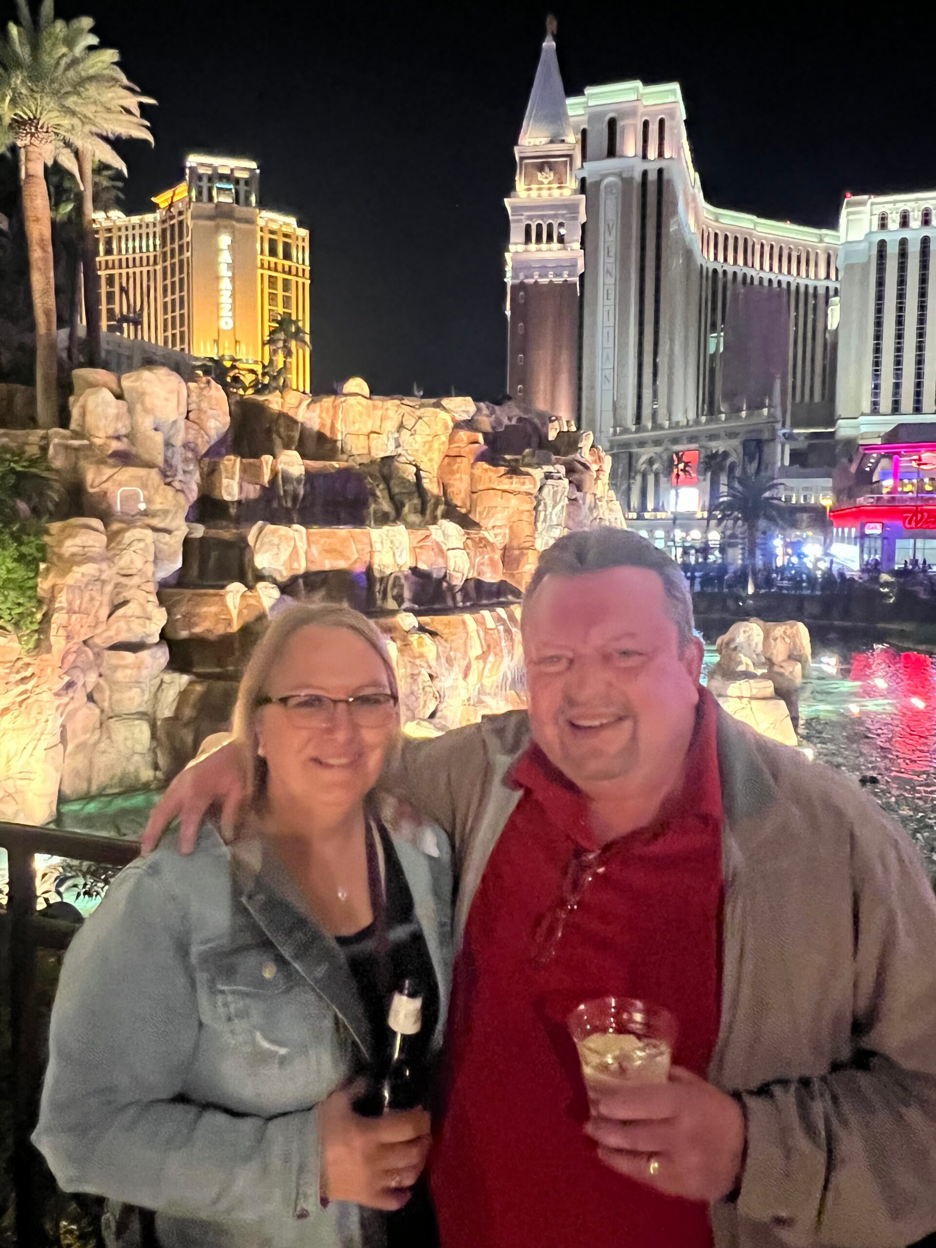 Smiling couple posing at night in Las Vegas, holding drinks, with neon-lit hotel towers and rock fountain behind them.