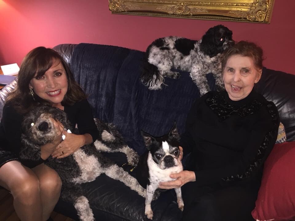 Two women on a blue sofa with three dogs: a fluffy black-and-white dog in the left woman's arms, a small bulldog on the right woman's lap, and another dog on the back cushion4.