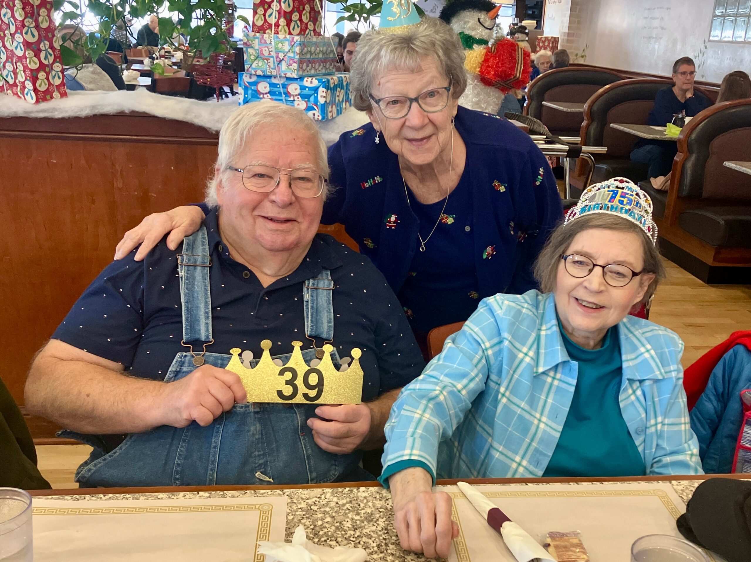 Two elderly friends pose for a birthday photo in a restaurant, with gifts and festive decorations in the background.