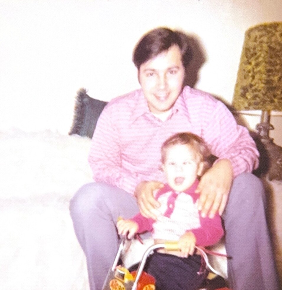 Man in pink striped shirt sitting on a couch with a toddler on a toy tricycle.