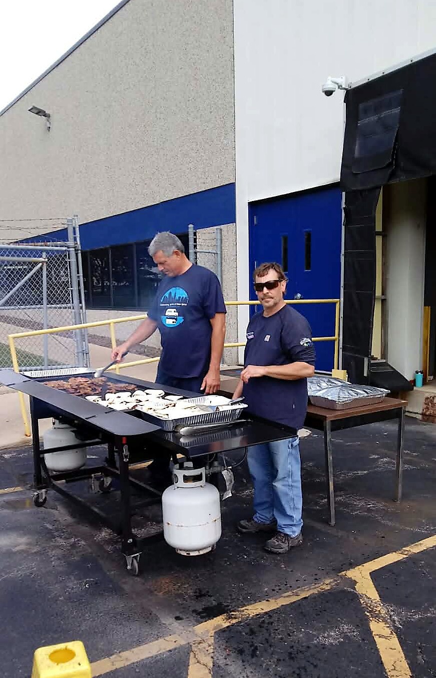 Two men cook on a large outdoor grill beside a warehouse, flip meat on the griddle using tongs; a propane tank sits beneath the grill.