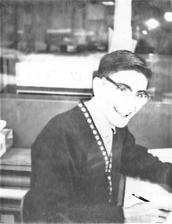 Smiling person with glasses seated at a desk, writing with a pencil in a black-and-white photo.