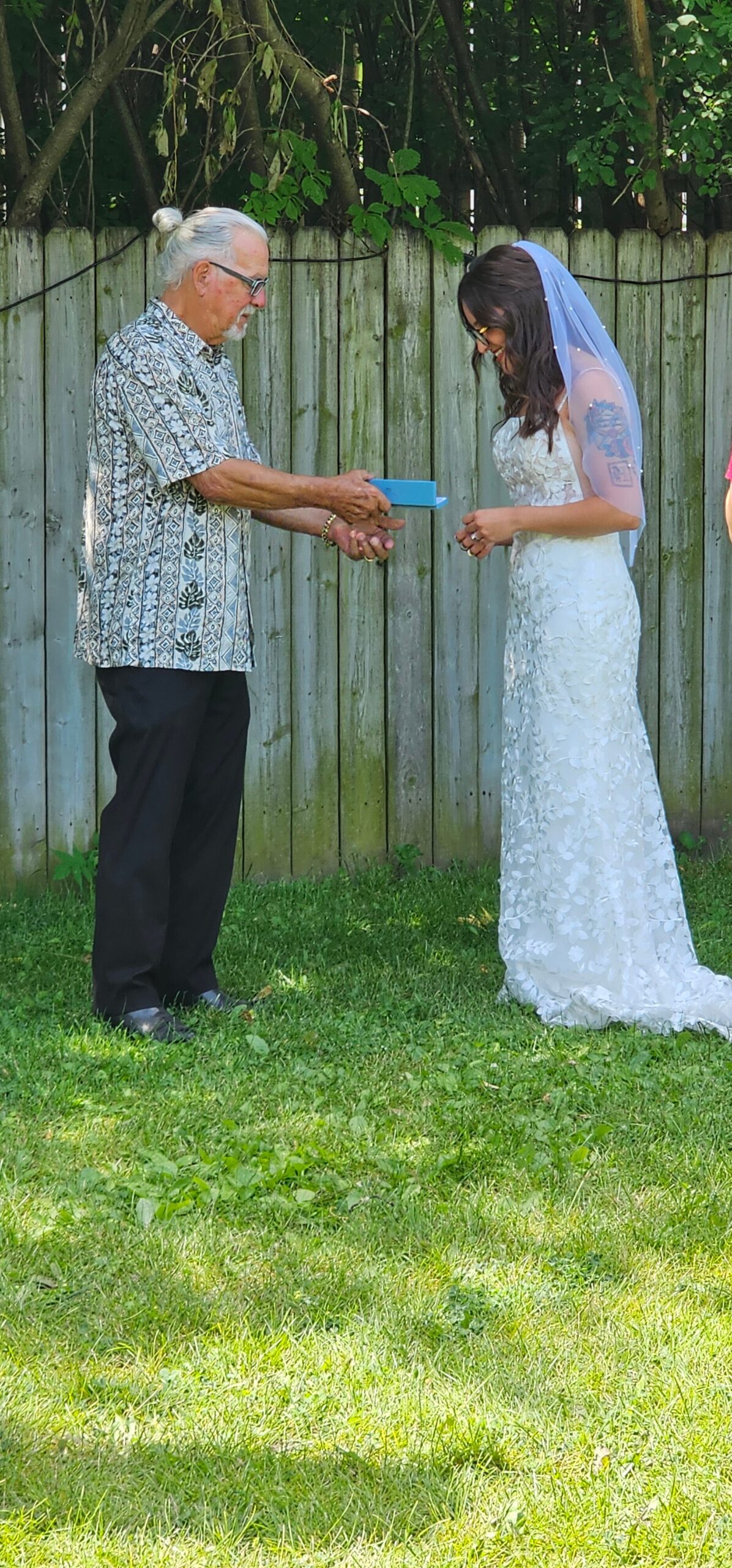 Bride in a lace wedding dress and veil receives a small blue box from an older man outdoors in a backyard, near a wooden fence.