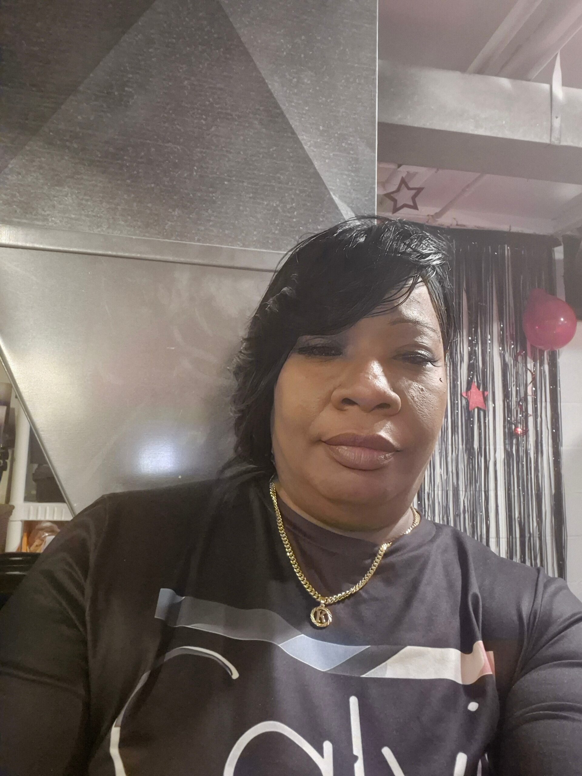 Close-up selfie of a smiling woman in a black graphic shirt and gold chain, with a metallic industrial backdrop and pink party decorations in the background.