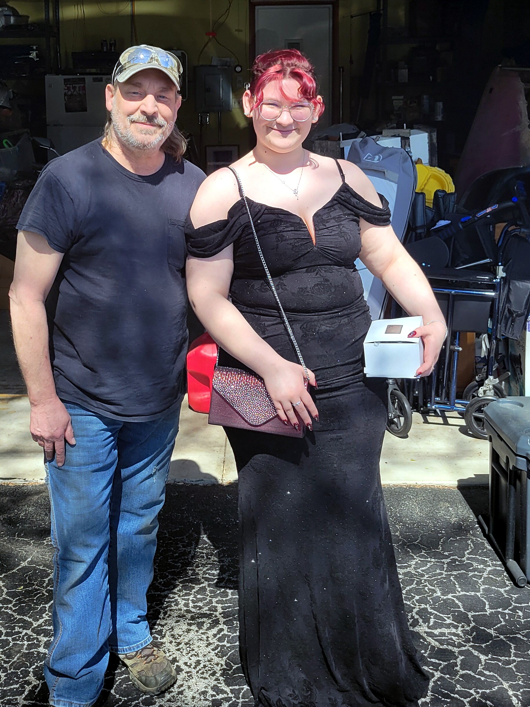 Man and woman pose together outside a workshop; man wears a cap and black T-shirt, woman in an off‑shoulder black gown holding a small white box and a sparkly purse.
