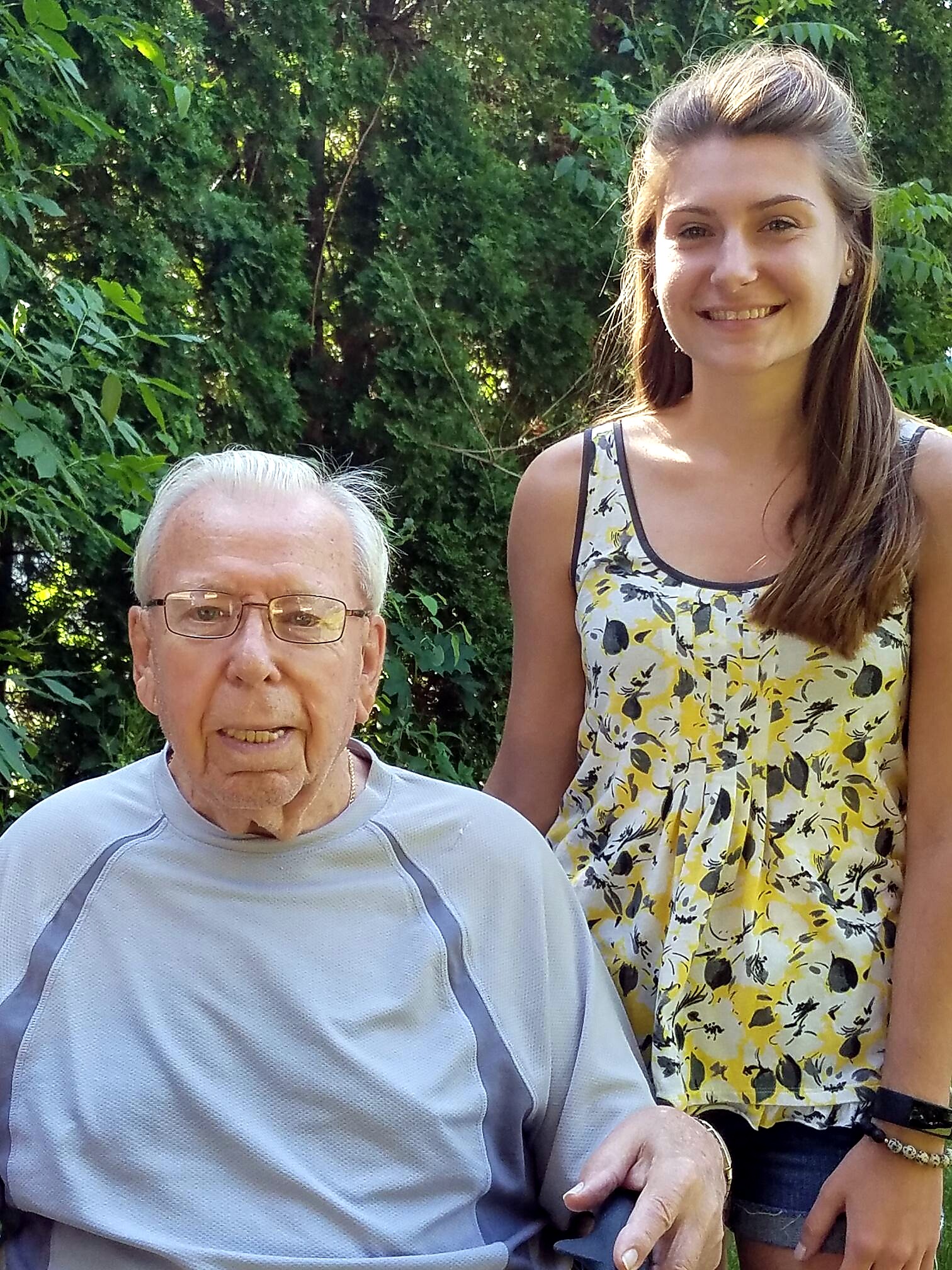 Smiling elderly man seated in foreground with a young woman standing beside him outdoors, green bushes in the background.
