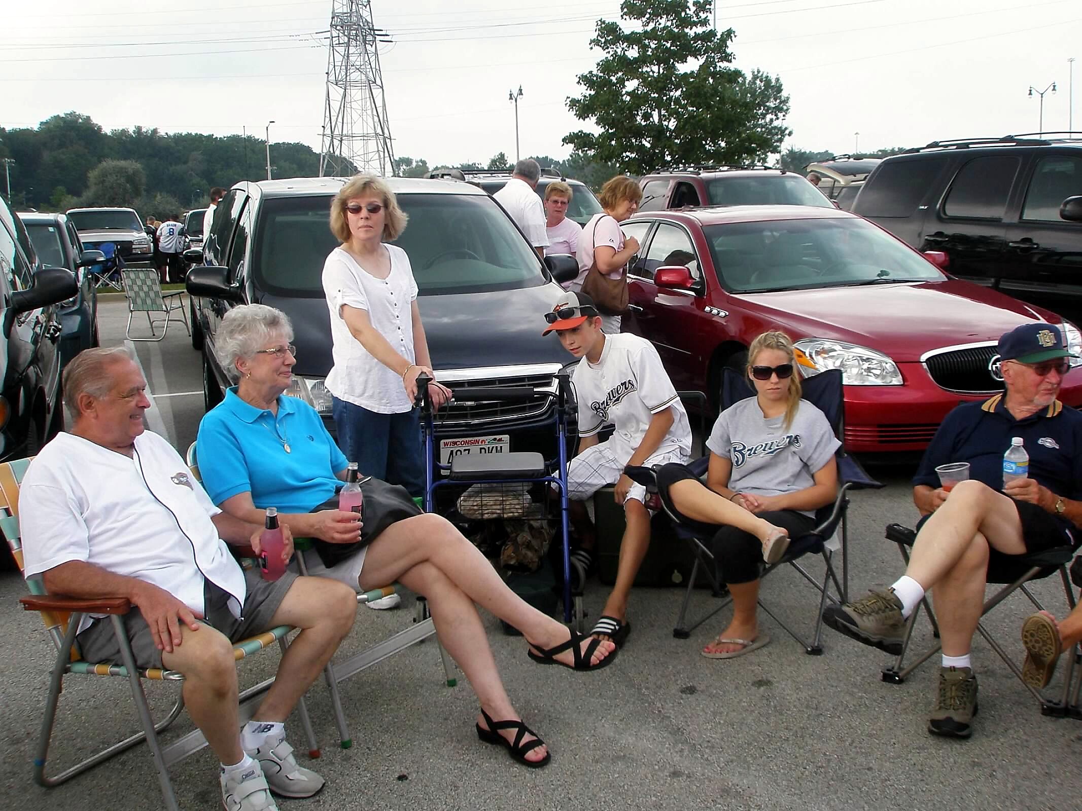 People relaxing in folding chairs in a crowded parking lot with many cars around.