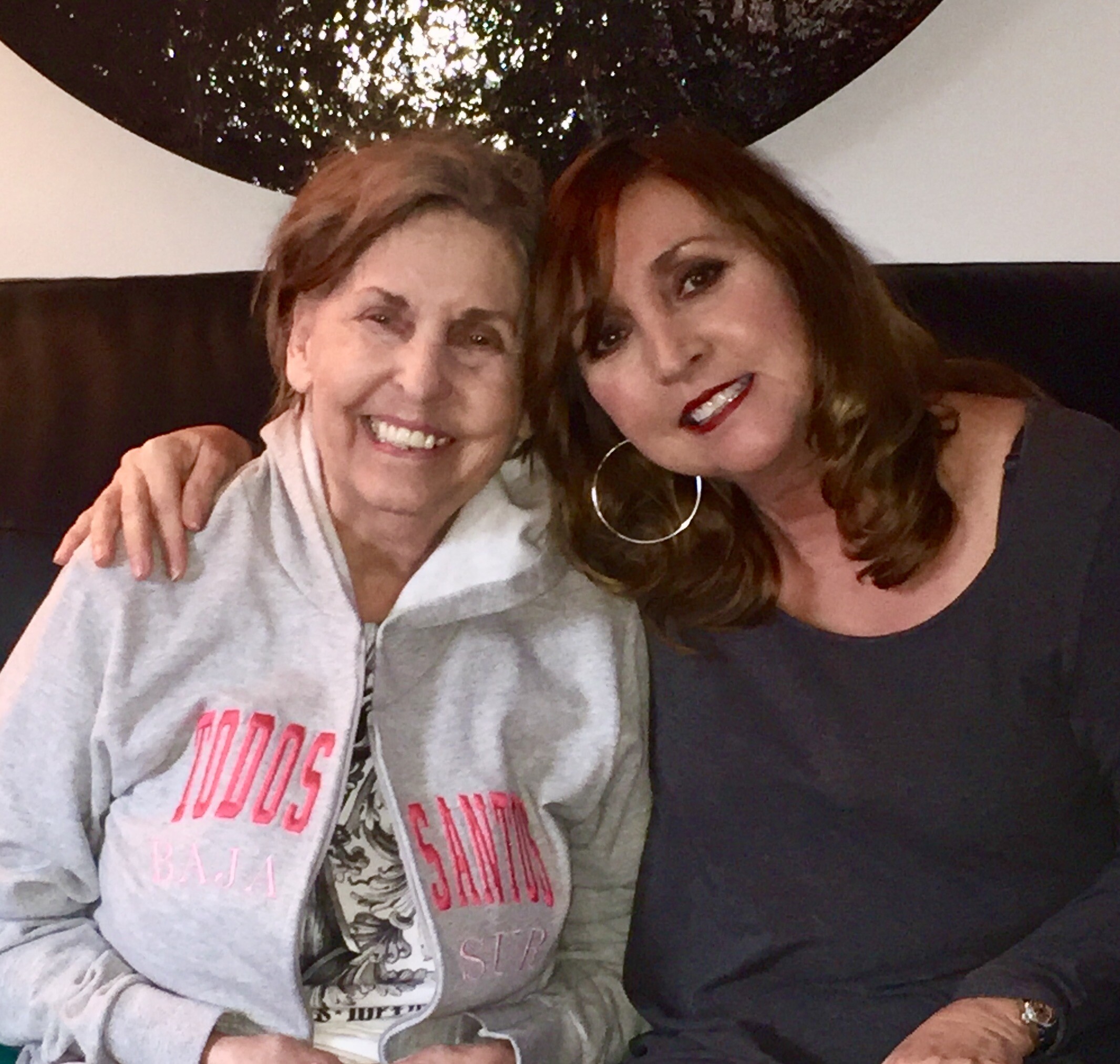 Two women posing for a selfie: an older woman with a gray hoodie on the left and a younger woman with hoop earrings and red lipstick on the right, smiling at the camera.