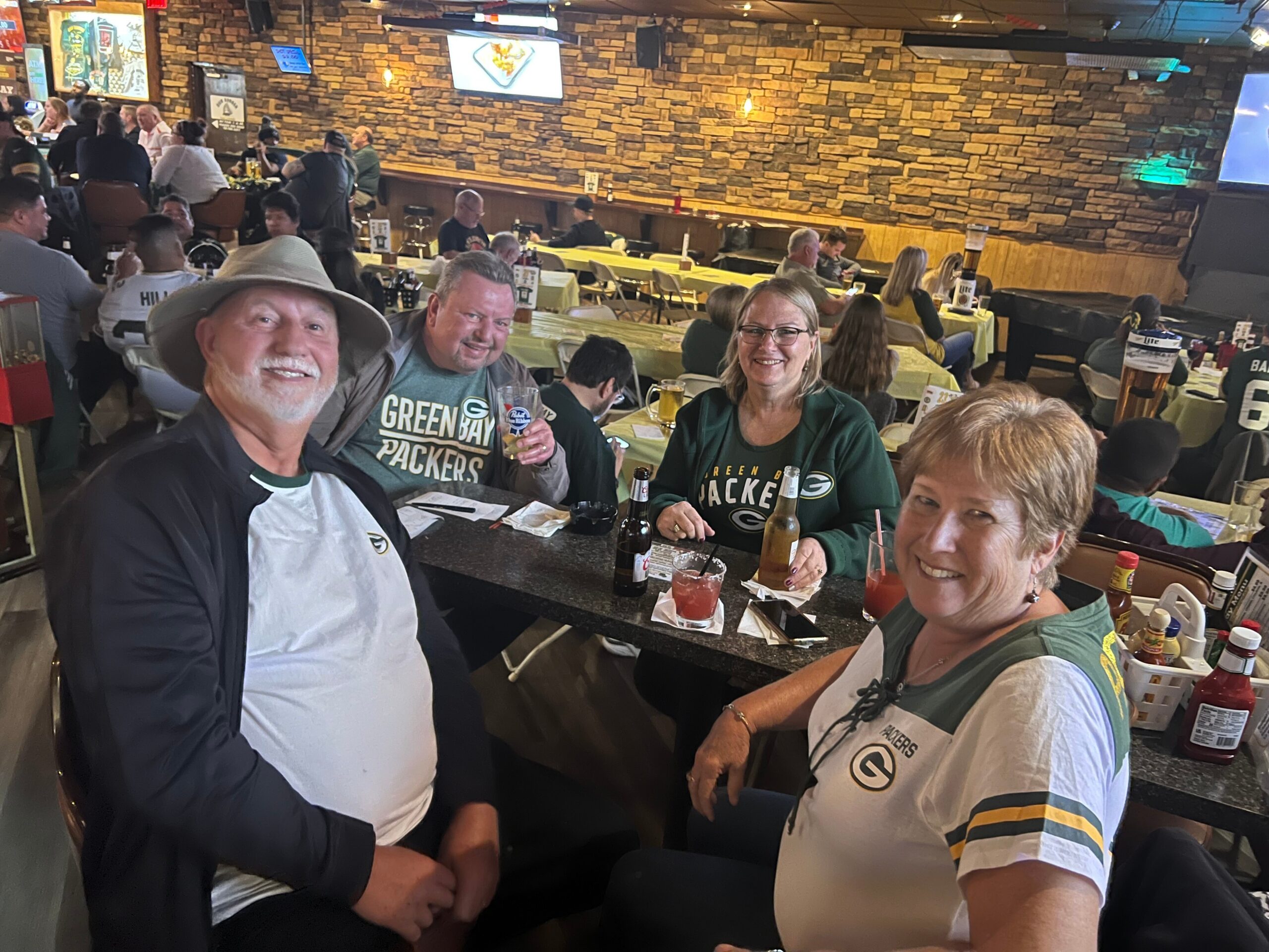 Group of Green Bay Packers fans posing for a selfie at a lively restaurant bar with drinks on the table and people in the background.