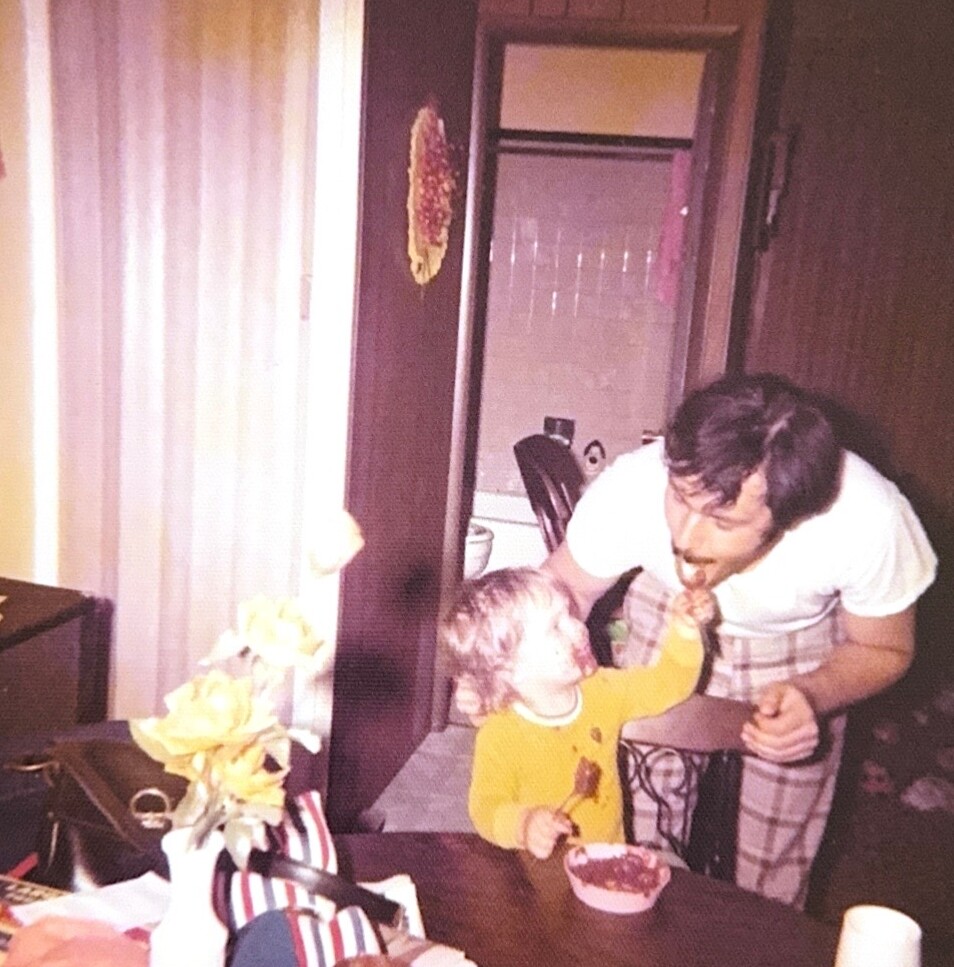 Man leaning over a dining table to feed a toddler with a spoon, indoors. The table has a vase of yellow flowers and clutter nearby.