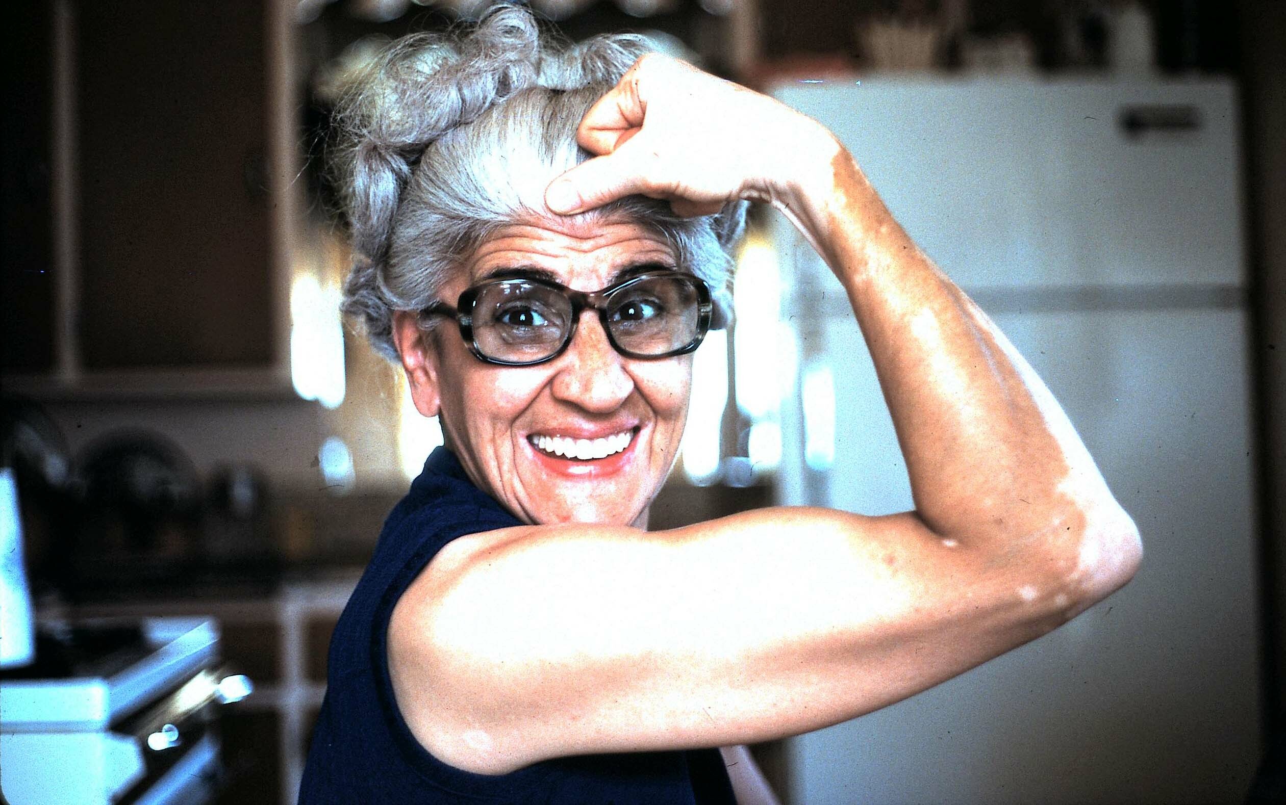 Older woman with gray hair and glasses smiling while tucking hair with both hands in a kitchen setting.