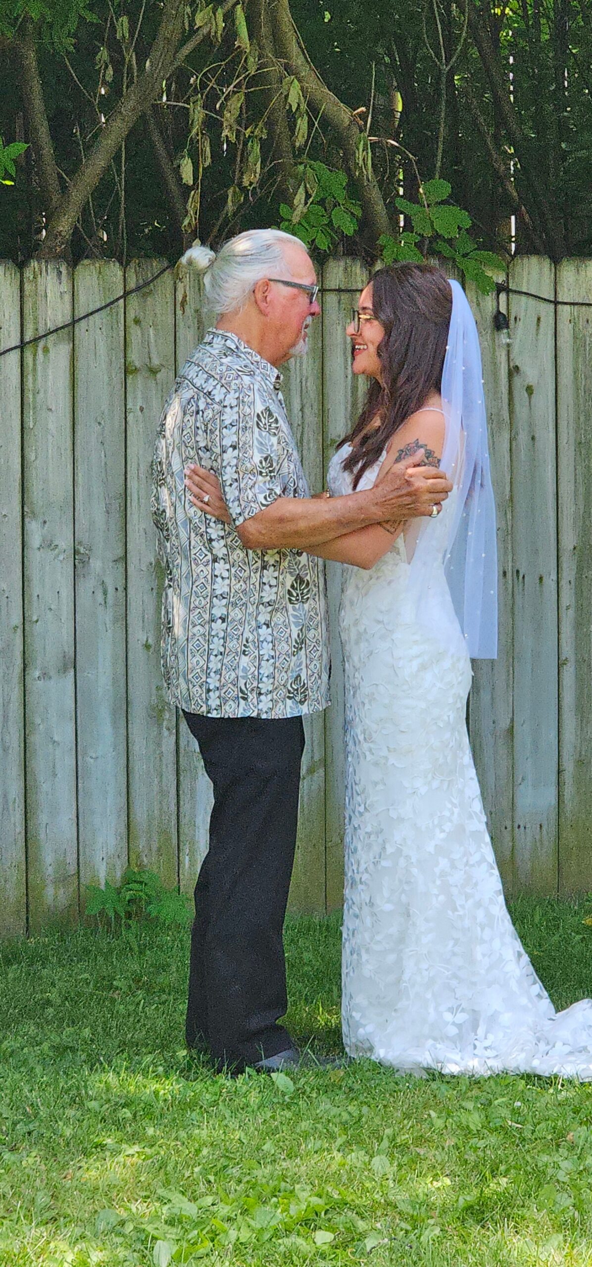 Bride in a white lace wedding dress and veil embraces an older man in a patterned shirt outdoors by a wooden fence.