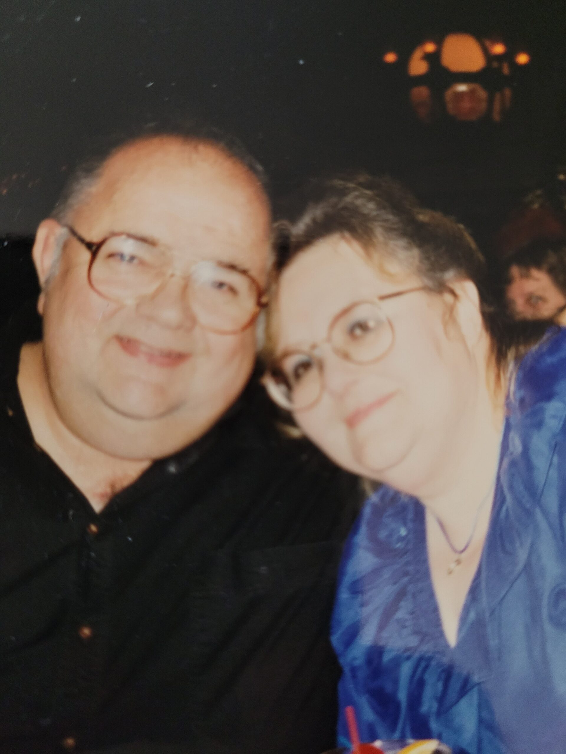 Two people posing for a selfie at night: man with glasses in a black shirt and woman in a blue jacket and glasses.