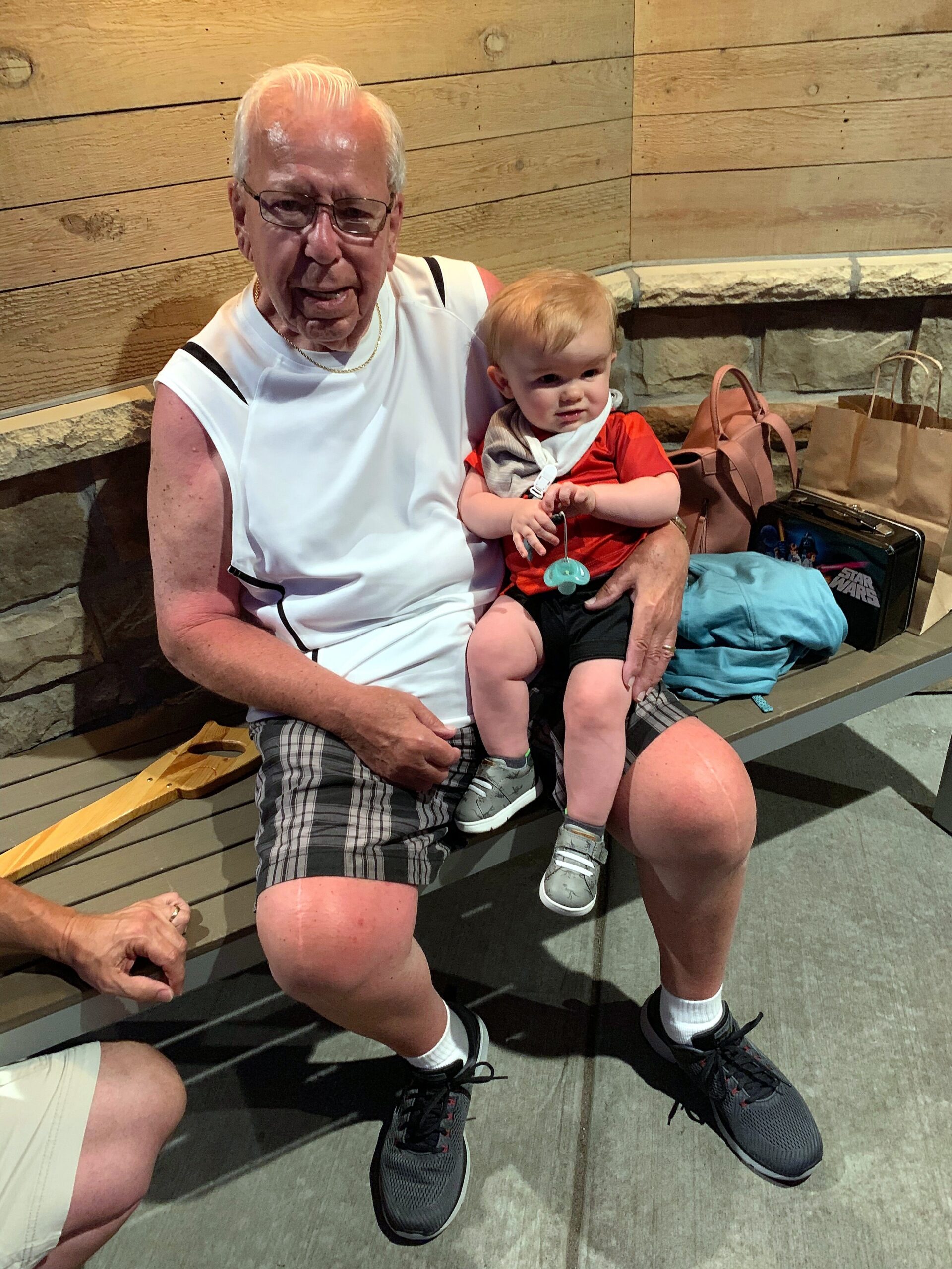 An elderly man with white hair and glasses sits on a bench, holding a small child in a red shirt as bags rest nearby.