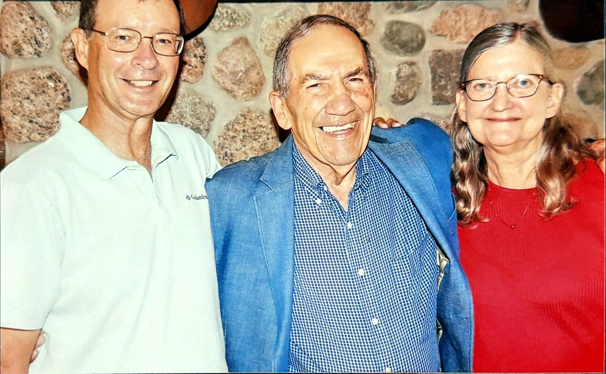 Three people posing together and smiling in front of a stone wall; man left in light shirt, center man in blue blazer, woman right in red with glasses.