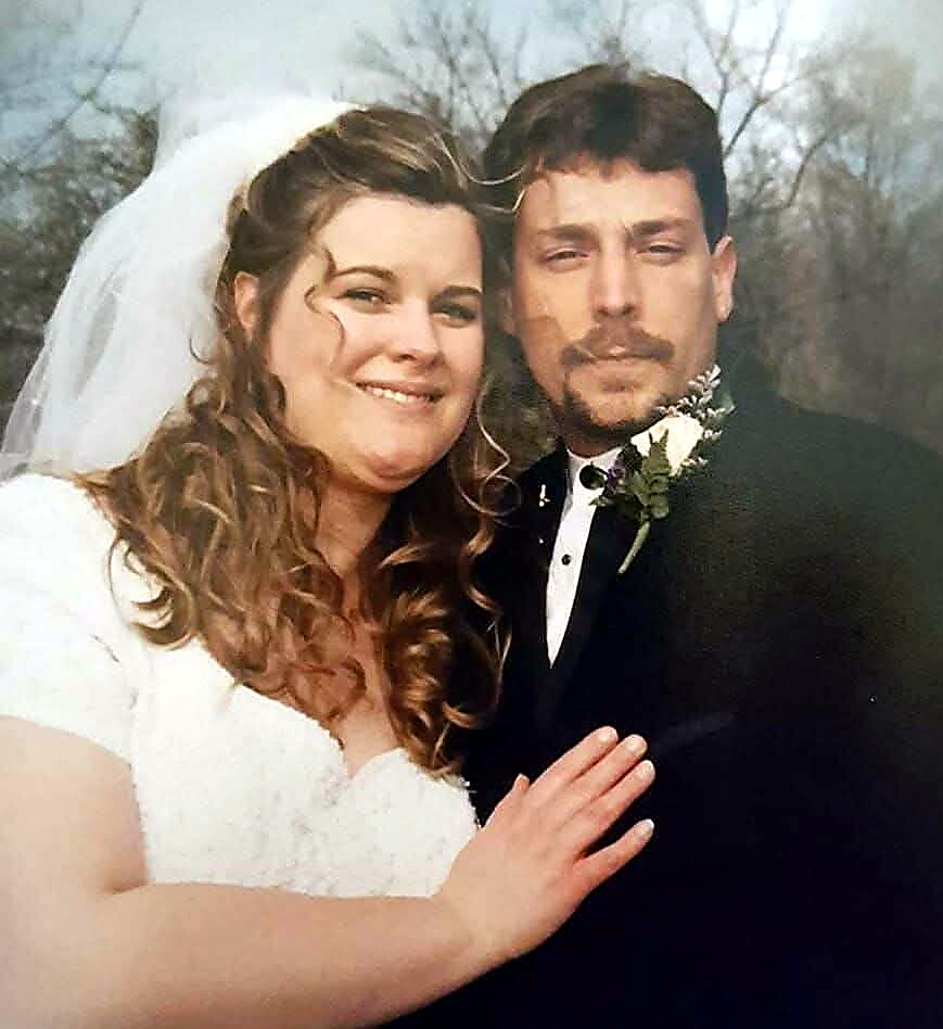 Smiling bride in a white wedding dress and veil stands with groom in a black suit outdoors on their wedding day.