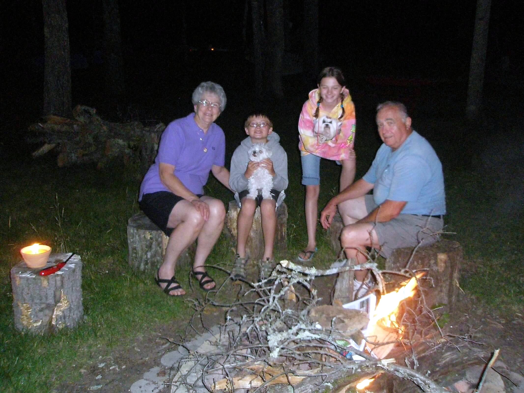A family of five sitting around a campfire at night in a wooded backyard, smiling at the camera with a small white dog.