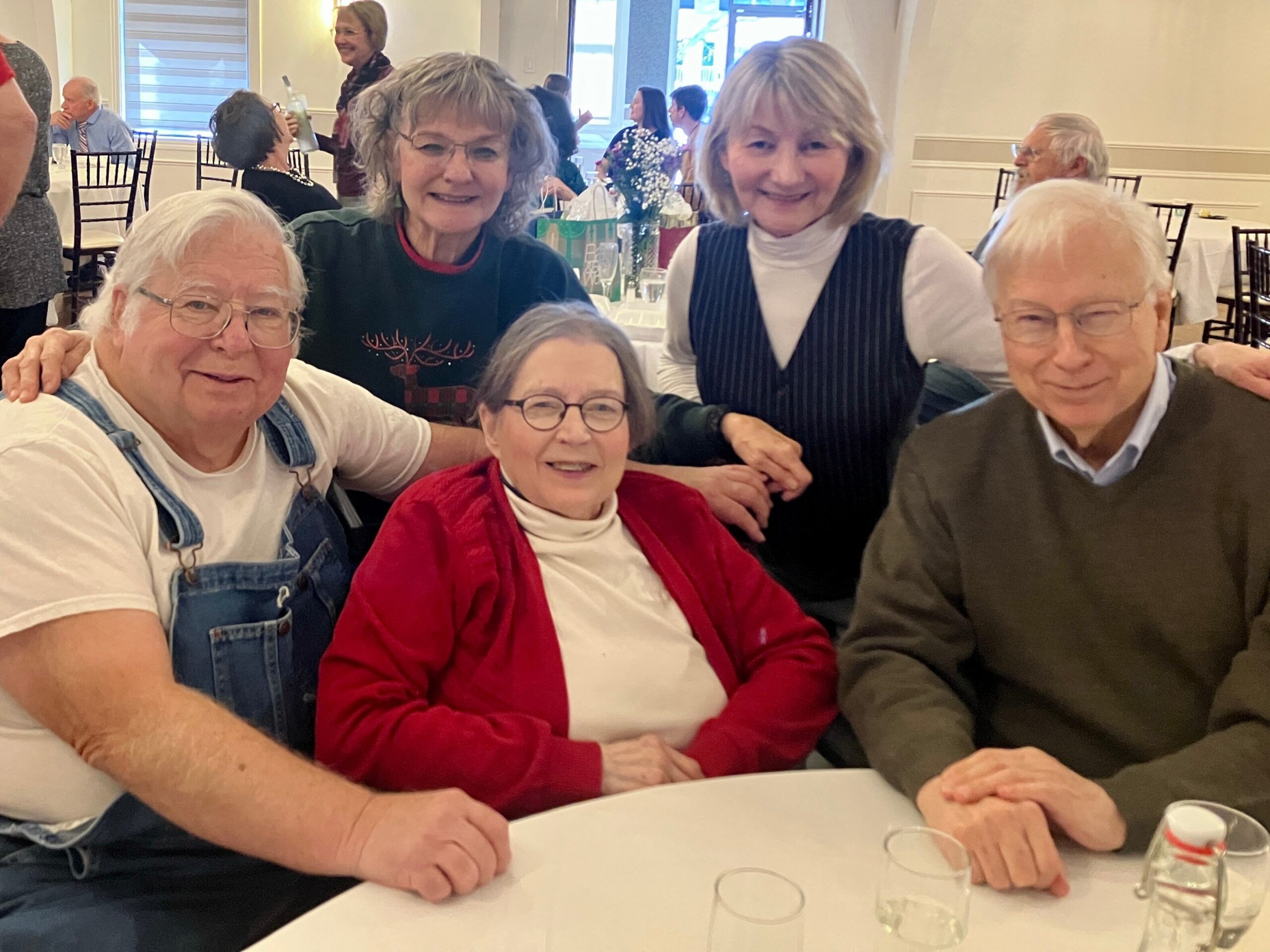 Group of smiling older adults gathered around a white-clothed table at a social event, with drinks in plastic cups and a small centerpiece visible in the background.