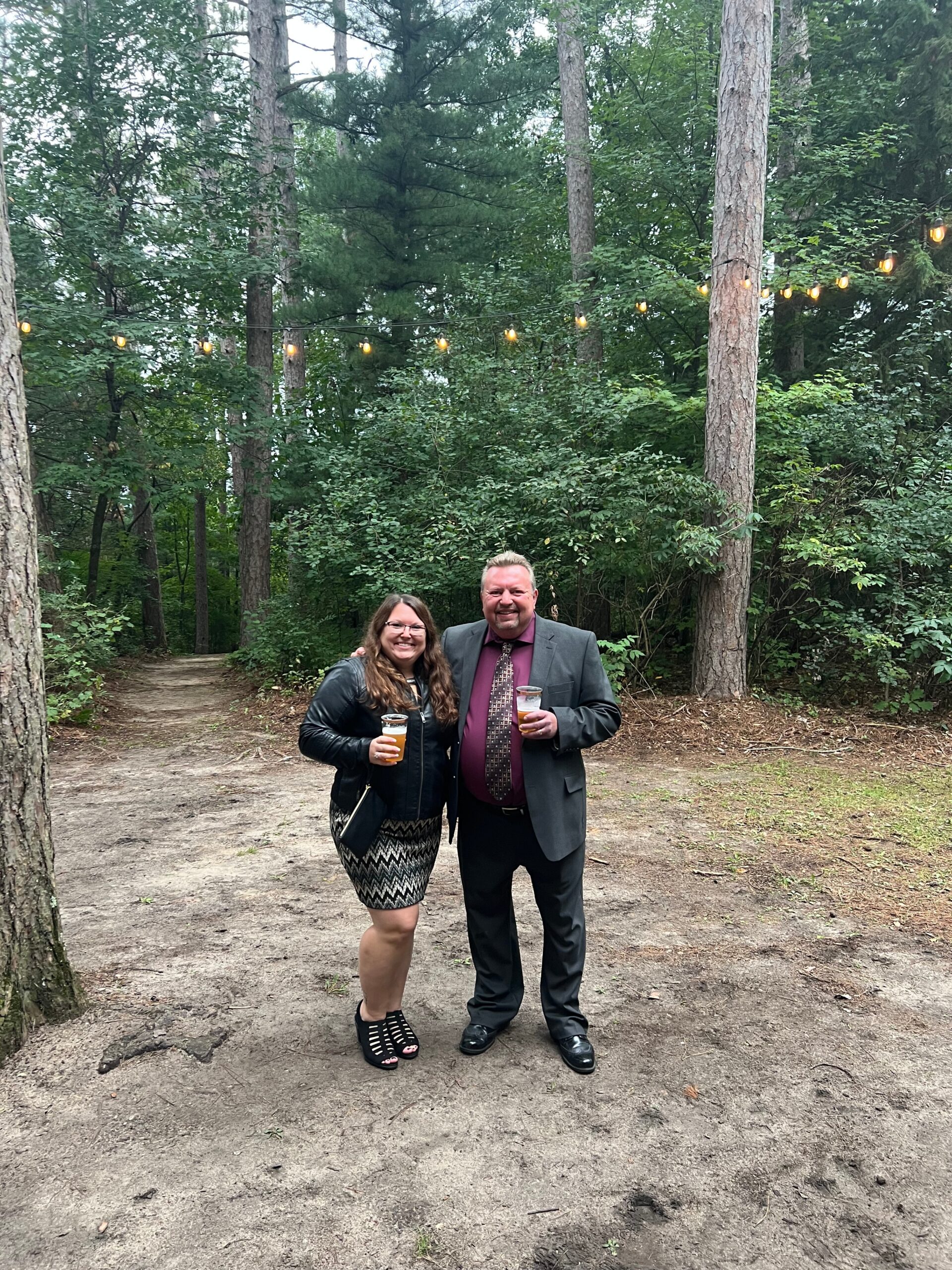 Smiling couple in a wooded area posing for a photo with drinks under string lights between trees.