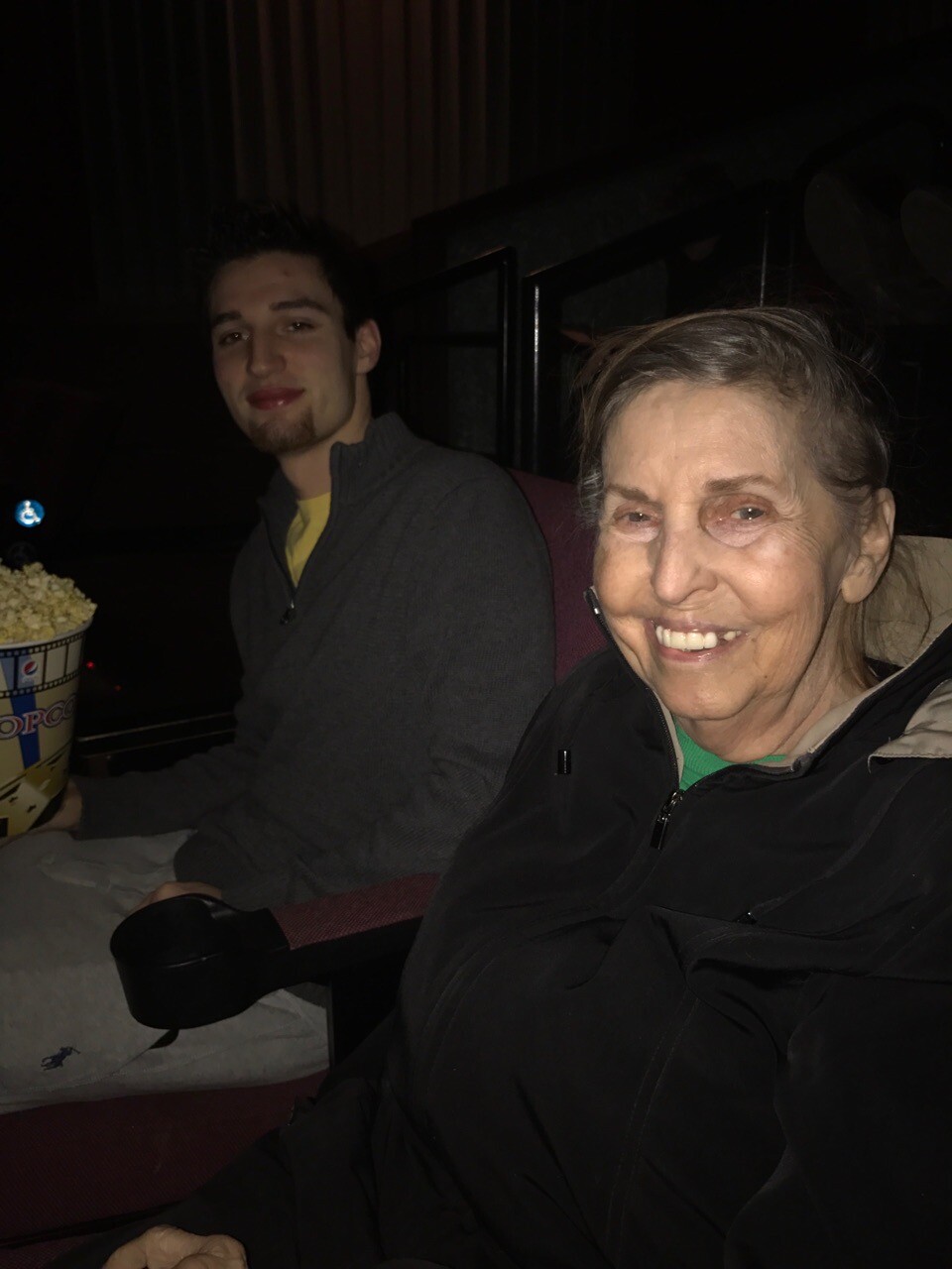 Smiling elderly woman in a dim movie theater beside a young man with a large popcorn bucket visible on the left.