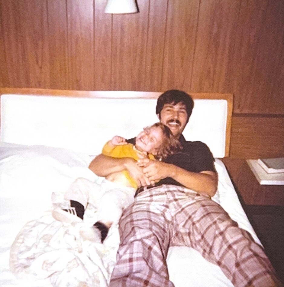 Man with a mustache lying in bed, hugging a smiling child in a yellow shirt under a plaid blanket in a wood-paneled room.